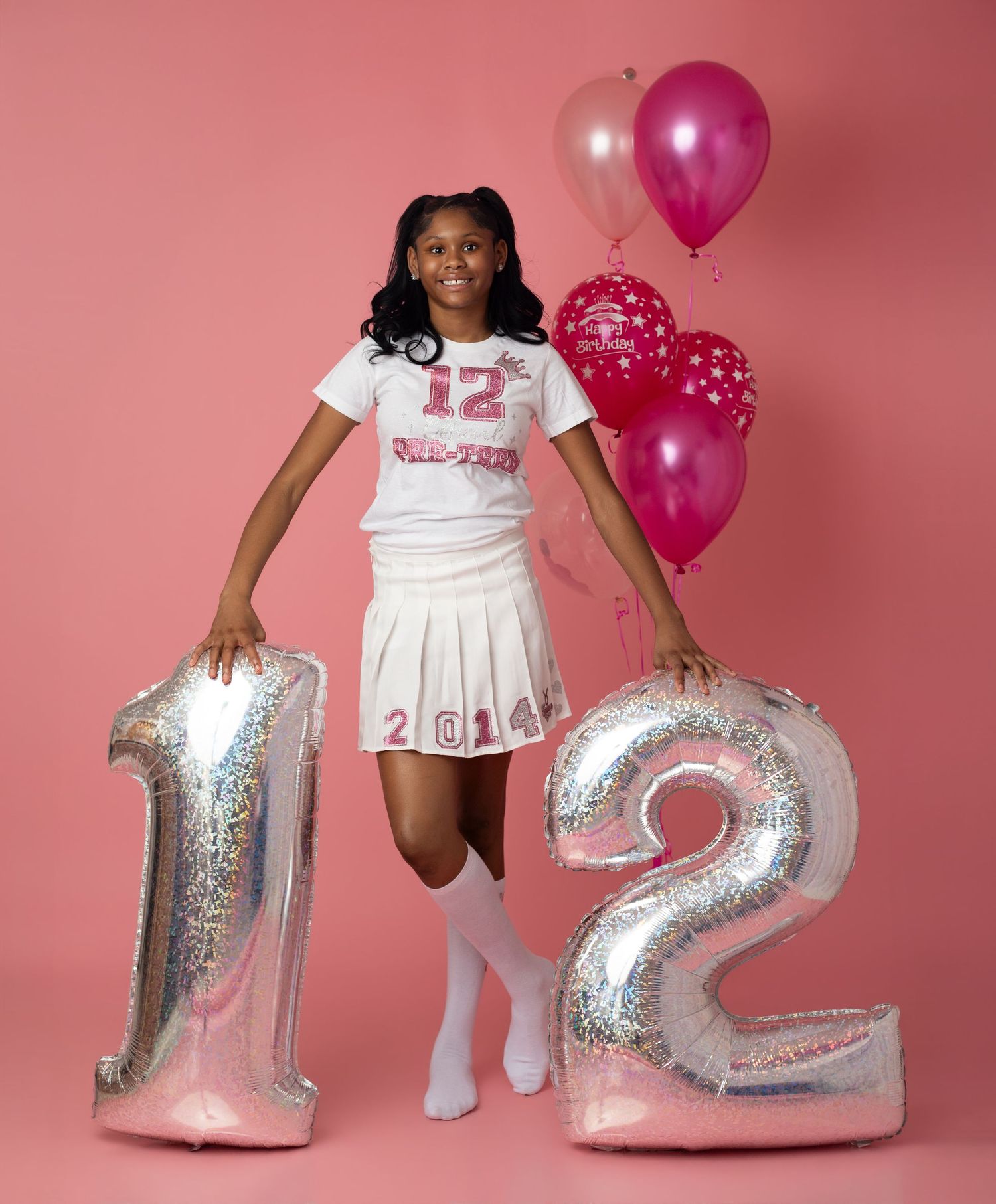 Girl in cheerleader uniform holding large number 12 balloons with pink balloons on a pink background.