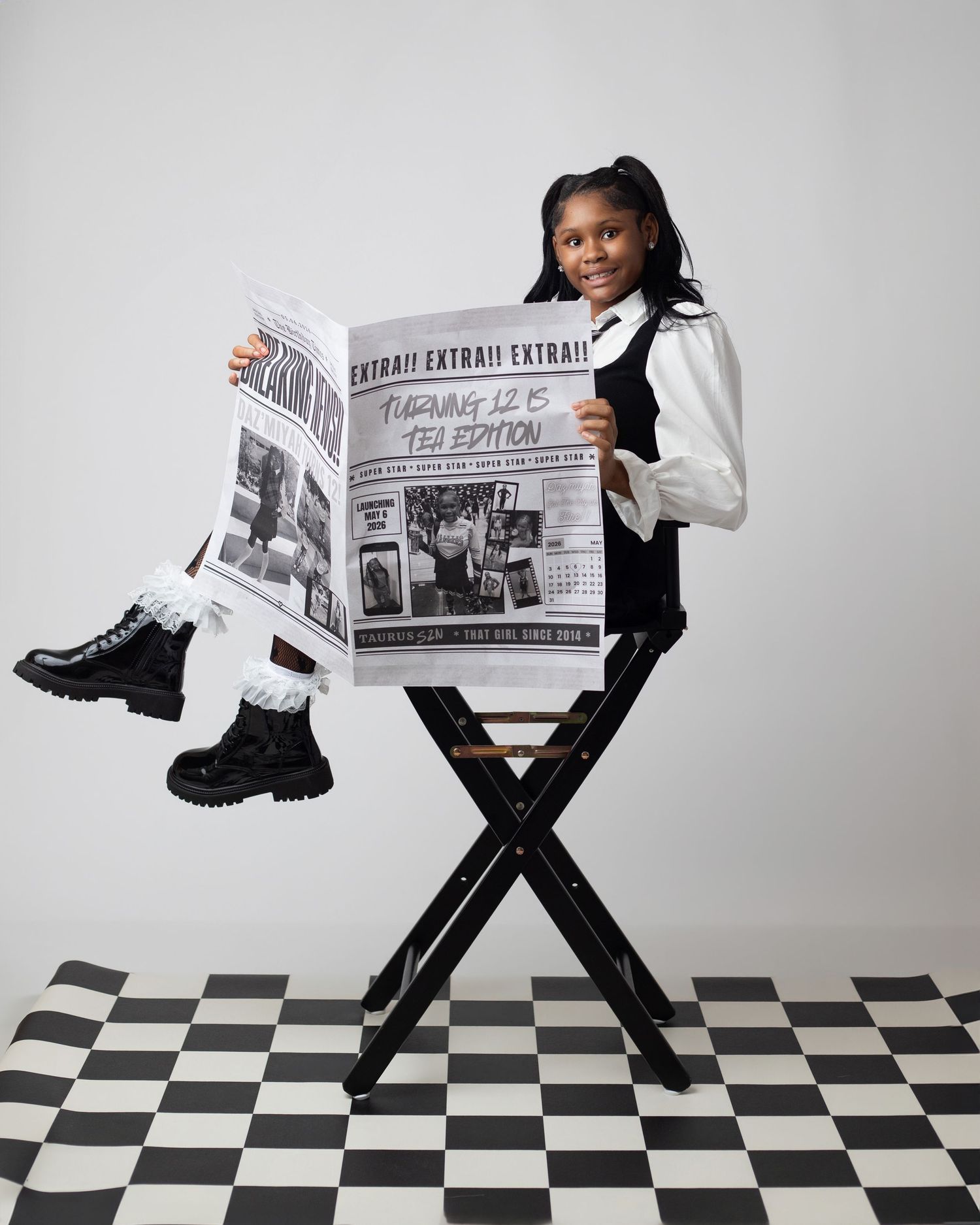 Young girl in black and white outfit sits on director's chair holding oversized newspaper on checkered floor.