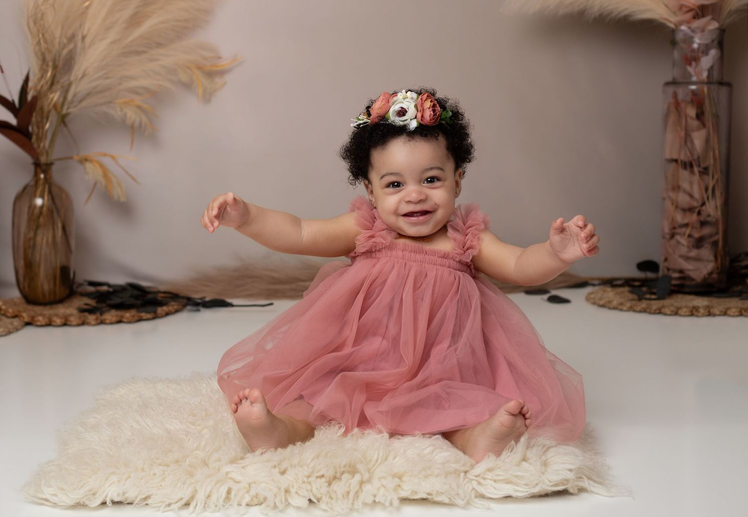 Smiling baby girl in pink tutu dress with floral headband sitting on white rug in professional photo studio.