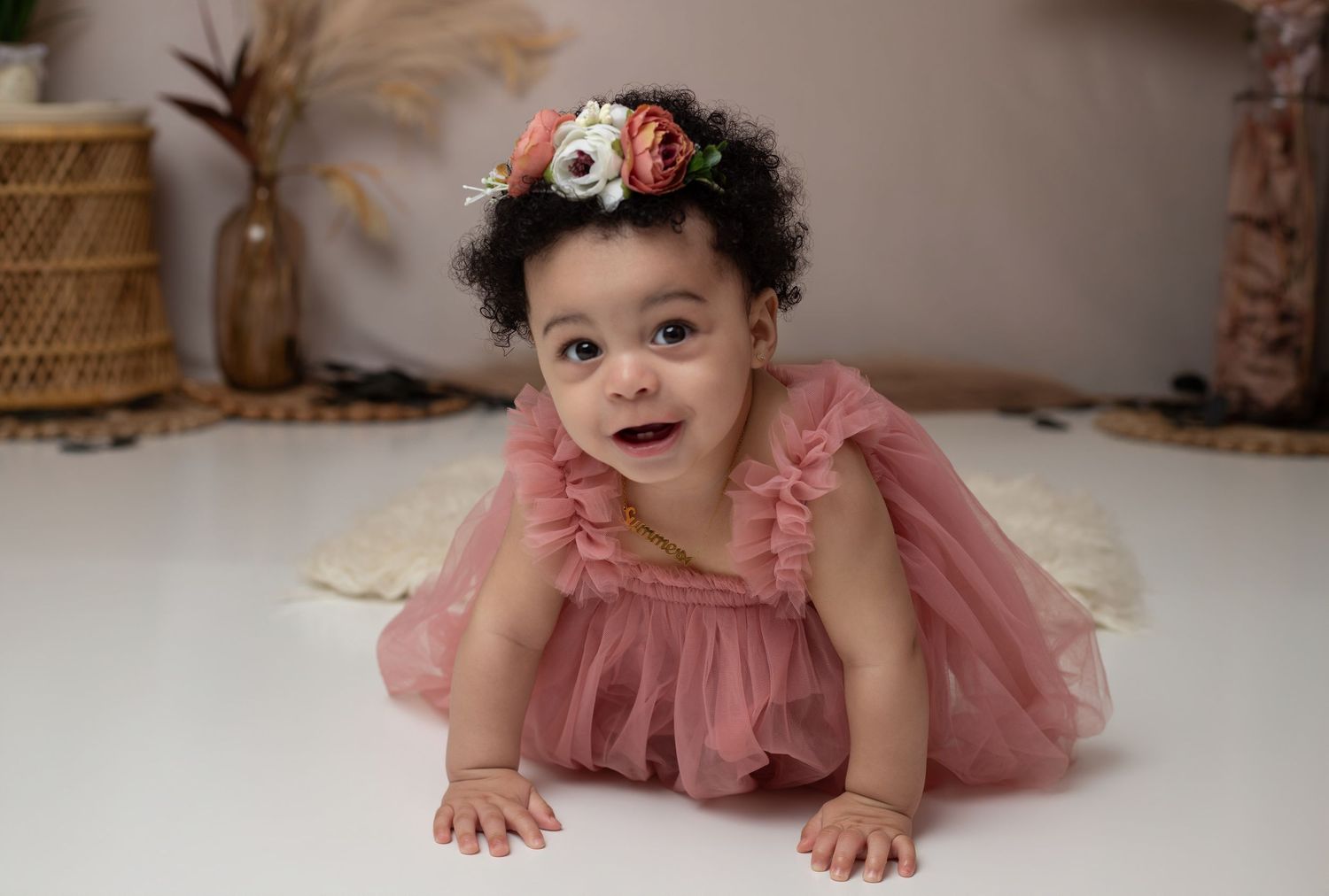Smiling baby girl in pink tulle dress and floral headband poses on white floor during first birthday photo shoot.