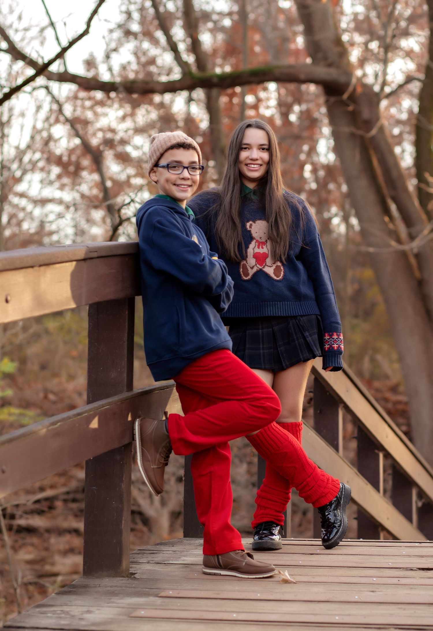 Two kids pose on a wooden bridge in navy sweaters, red pants, and plaid skirt, surrounded by autumn trees.