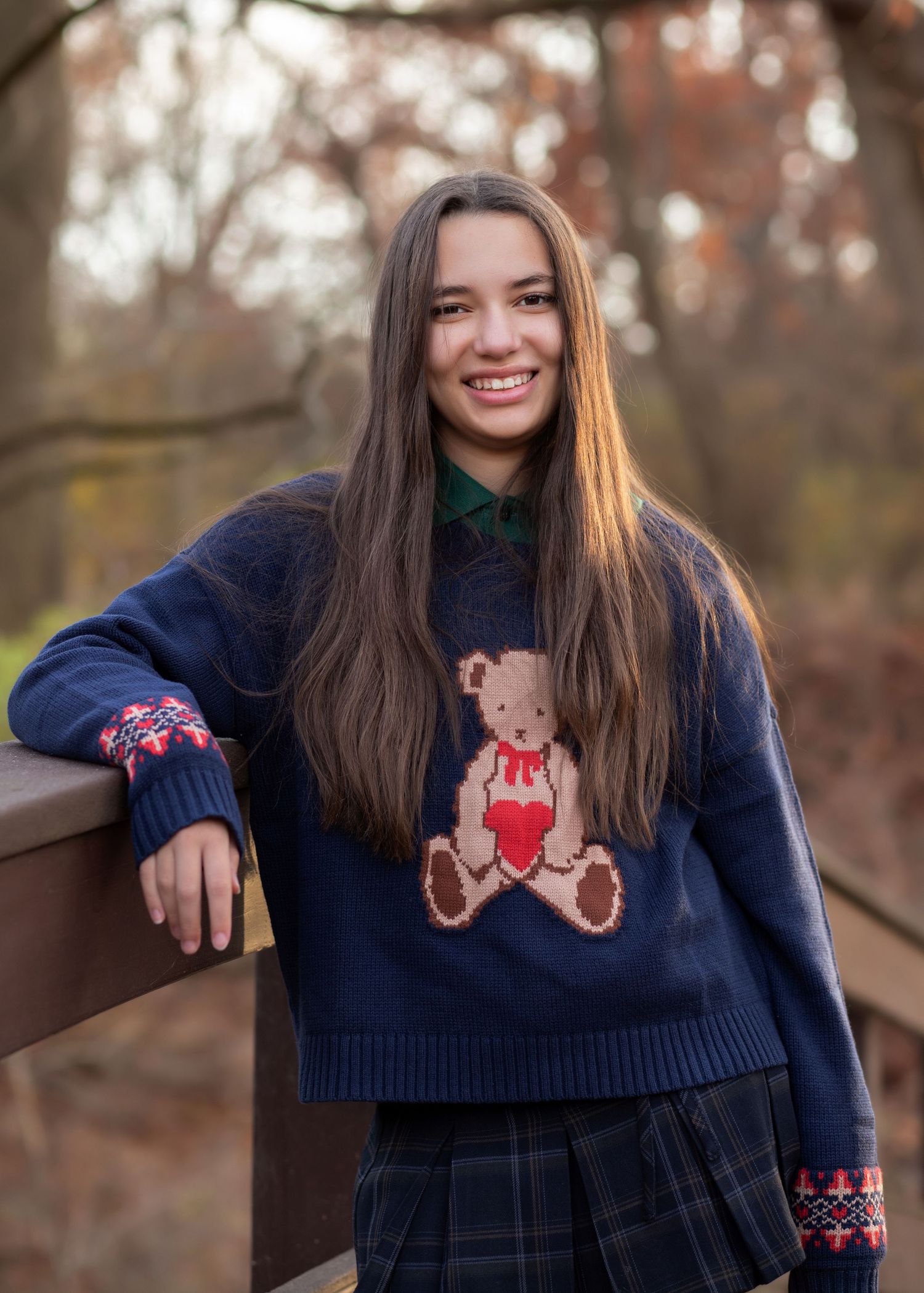 Smiling teenage girl in navy teddy bear sweater and plaid skirt leaning on wooden railing outdoors.