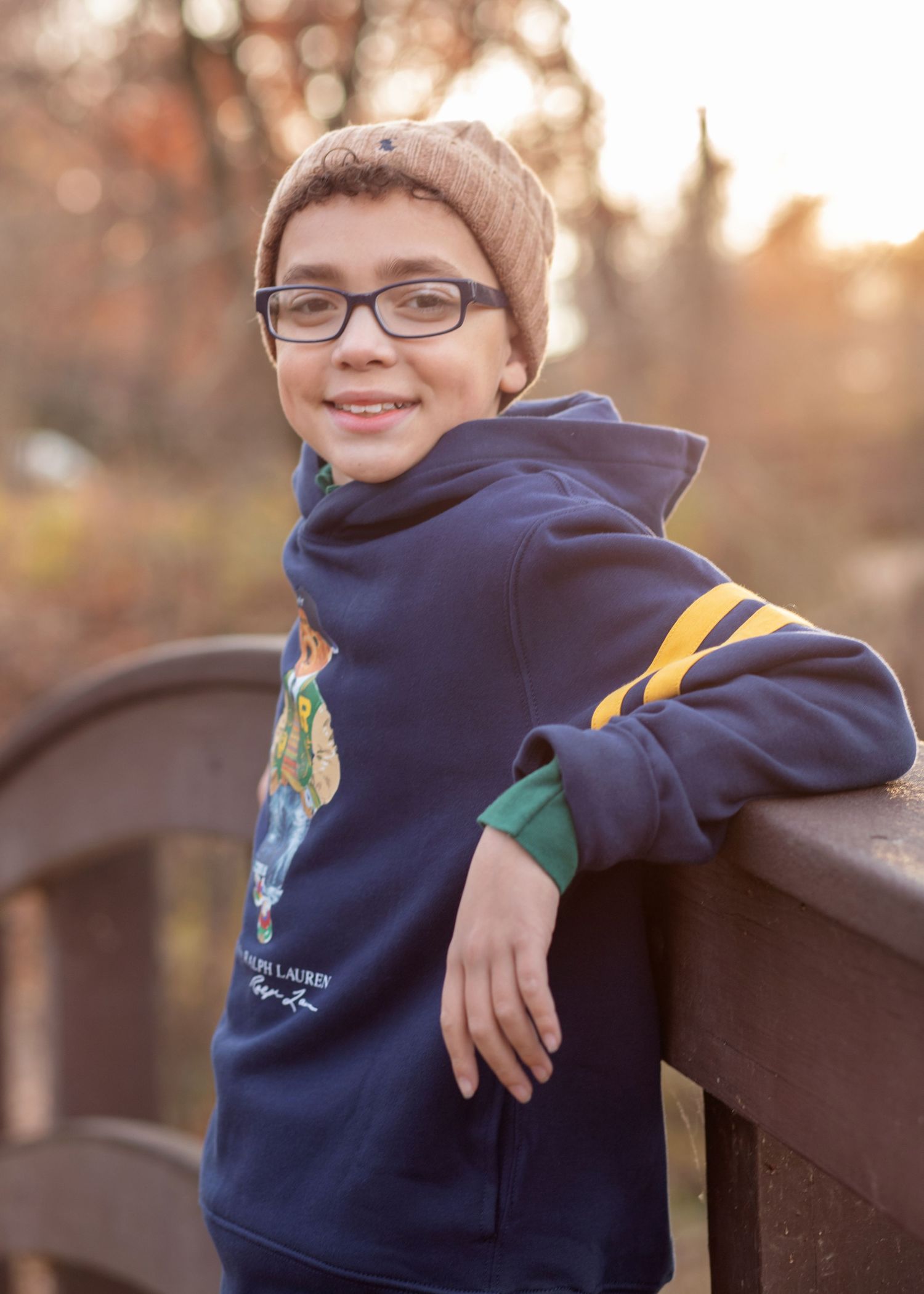 Smiling boy in navy Ralph Lauren hoodie and beanie glasses leaning on wooden bridge railing outdoors.