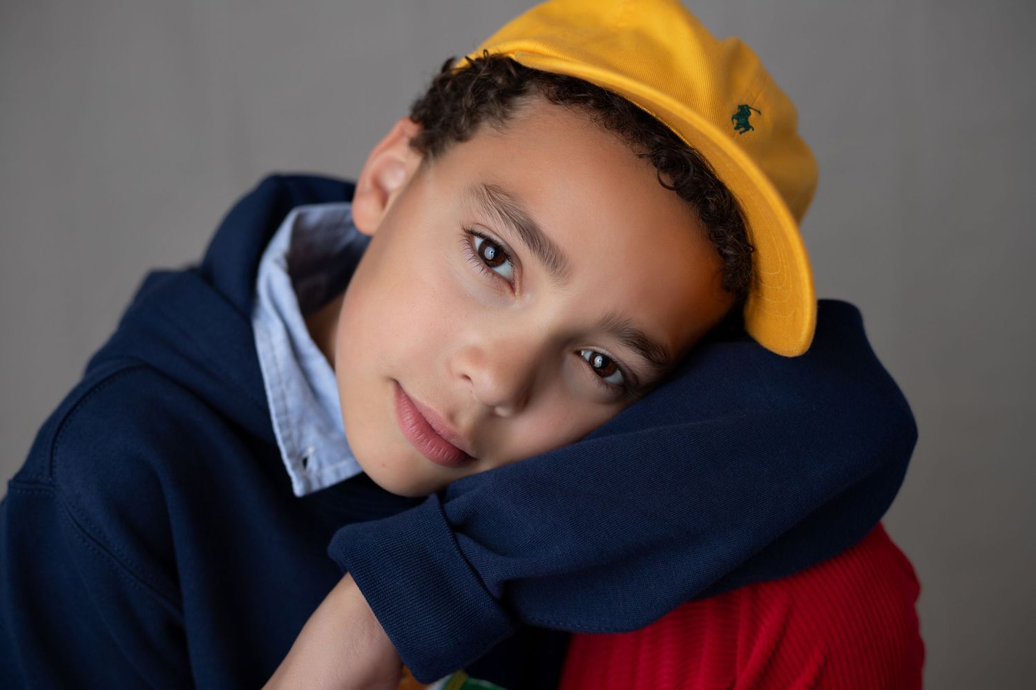 Young boy in yellow cap and blue hoodie resting chin on arms, looking at camera against gray background.