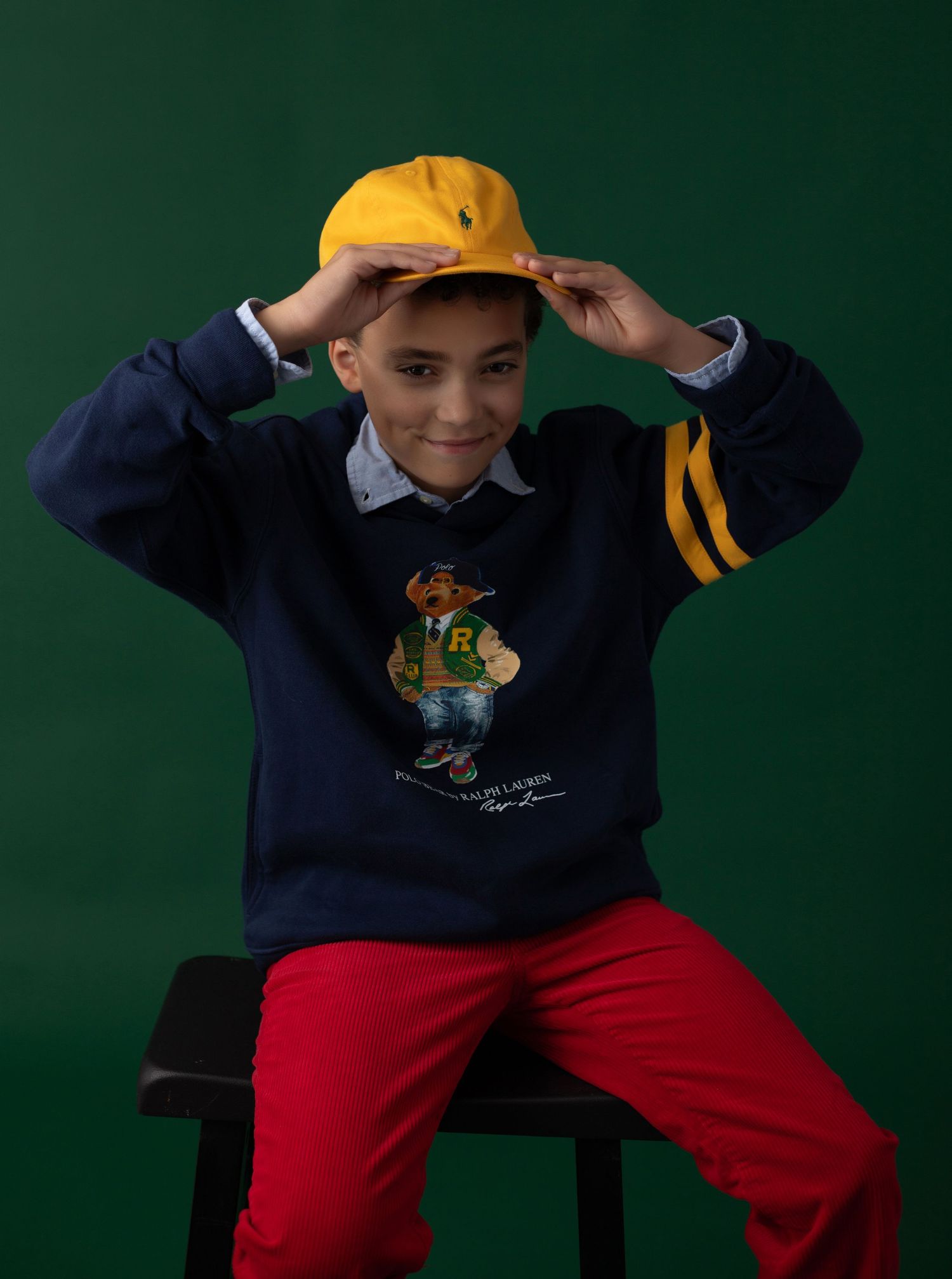 Boy wearing Ralph Lauren Polo Bear navy sweatshirt, yellow cap, and red pants, seated against green background.