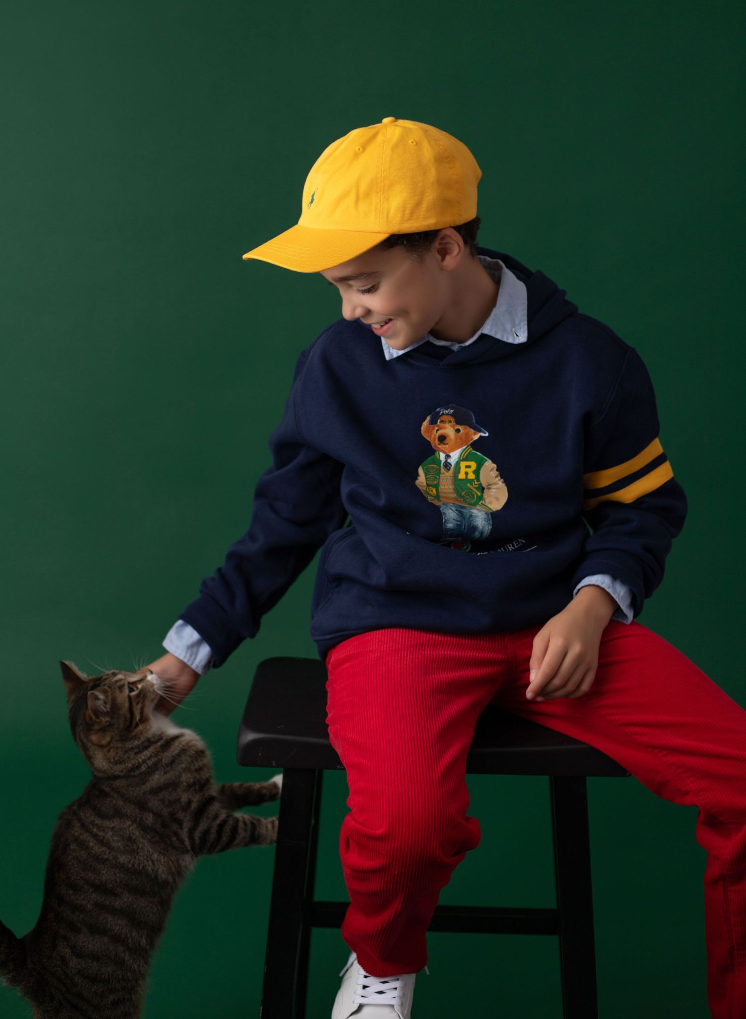 Boy in navy Ralph Lauren bear sweatshirt, yellow cap, and red pants sits on stool interacting with a tabby cat.