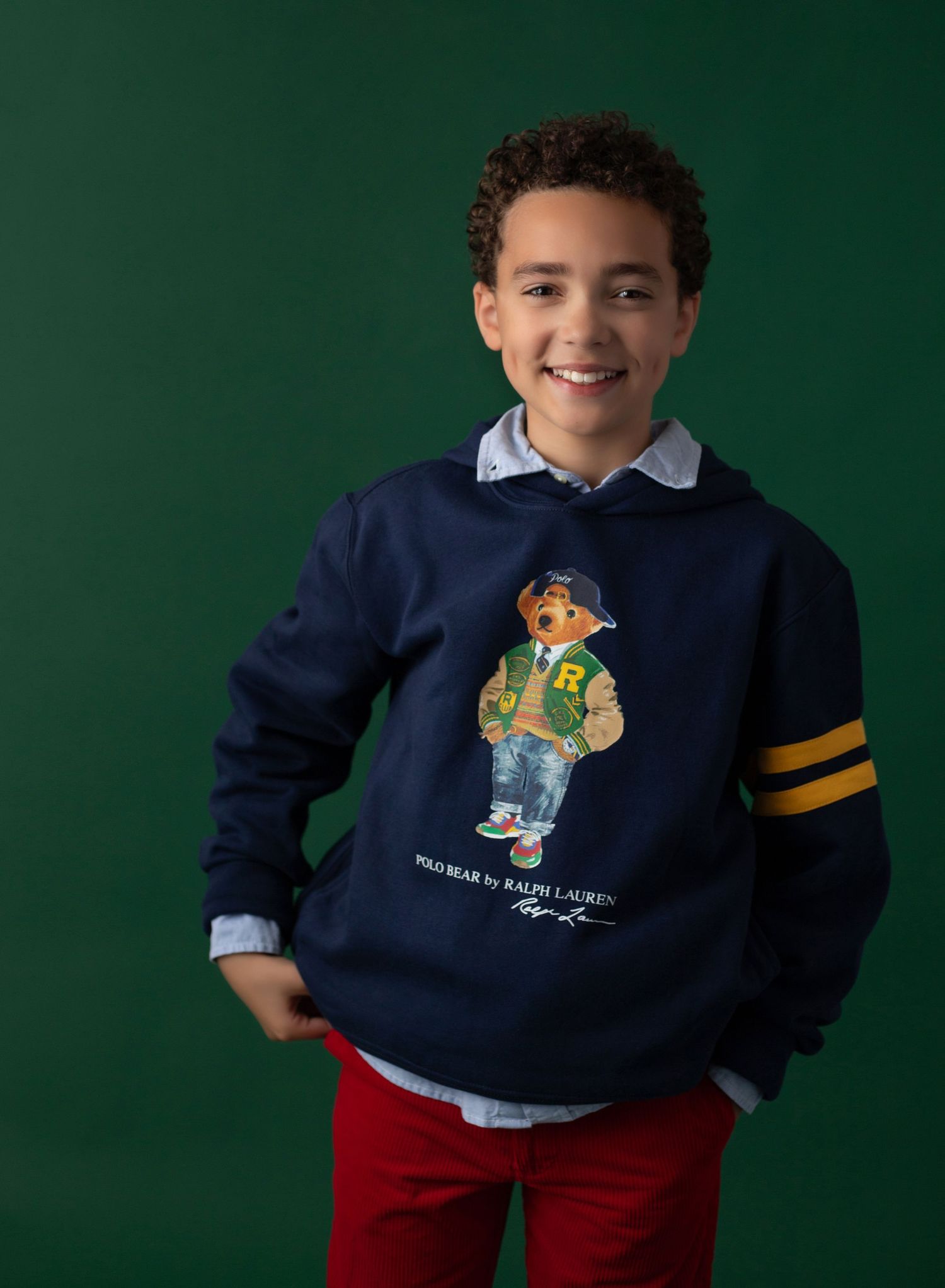 Boy wearing a navy Polo Bear by Ralph Lauren hoodie with red pants and a collared shirt, smiling against a green background.