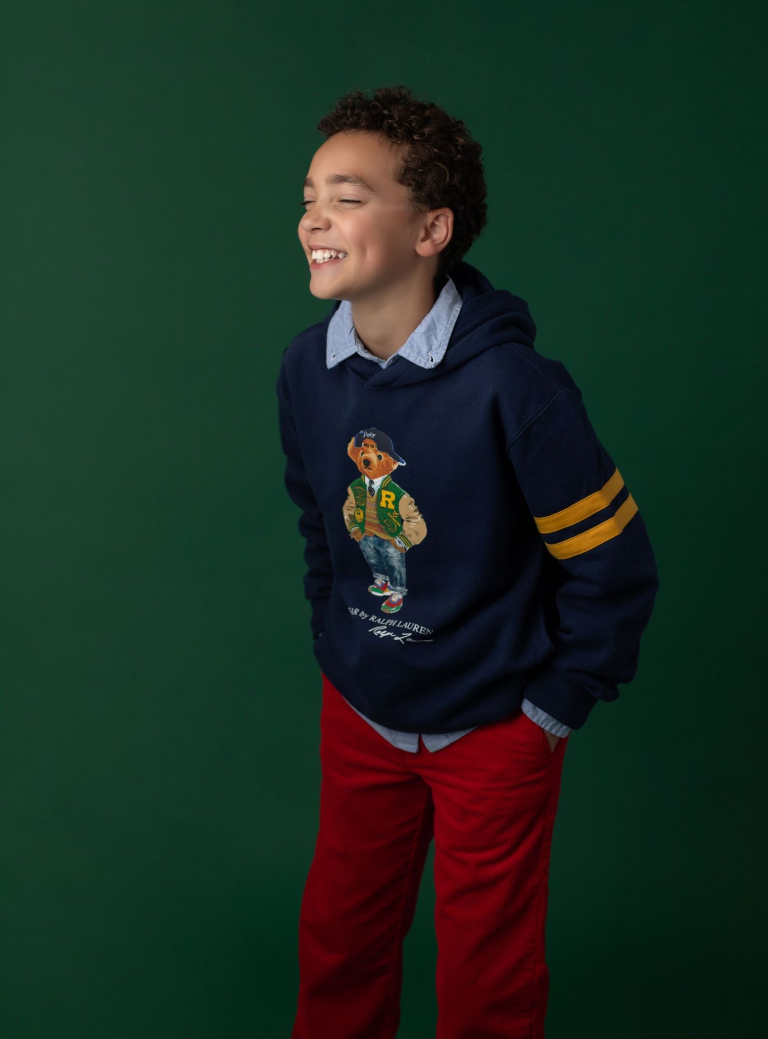 Boy smiling in navy Polo Ralph Lauren bear hoodie, blue shirt, and red pants against green background.