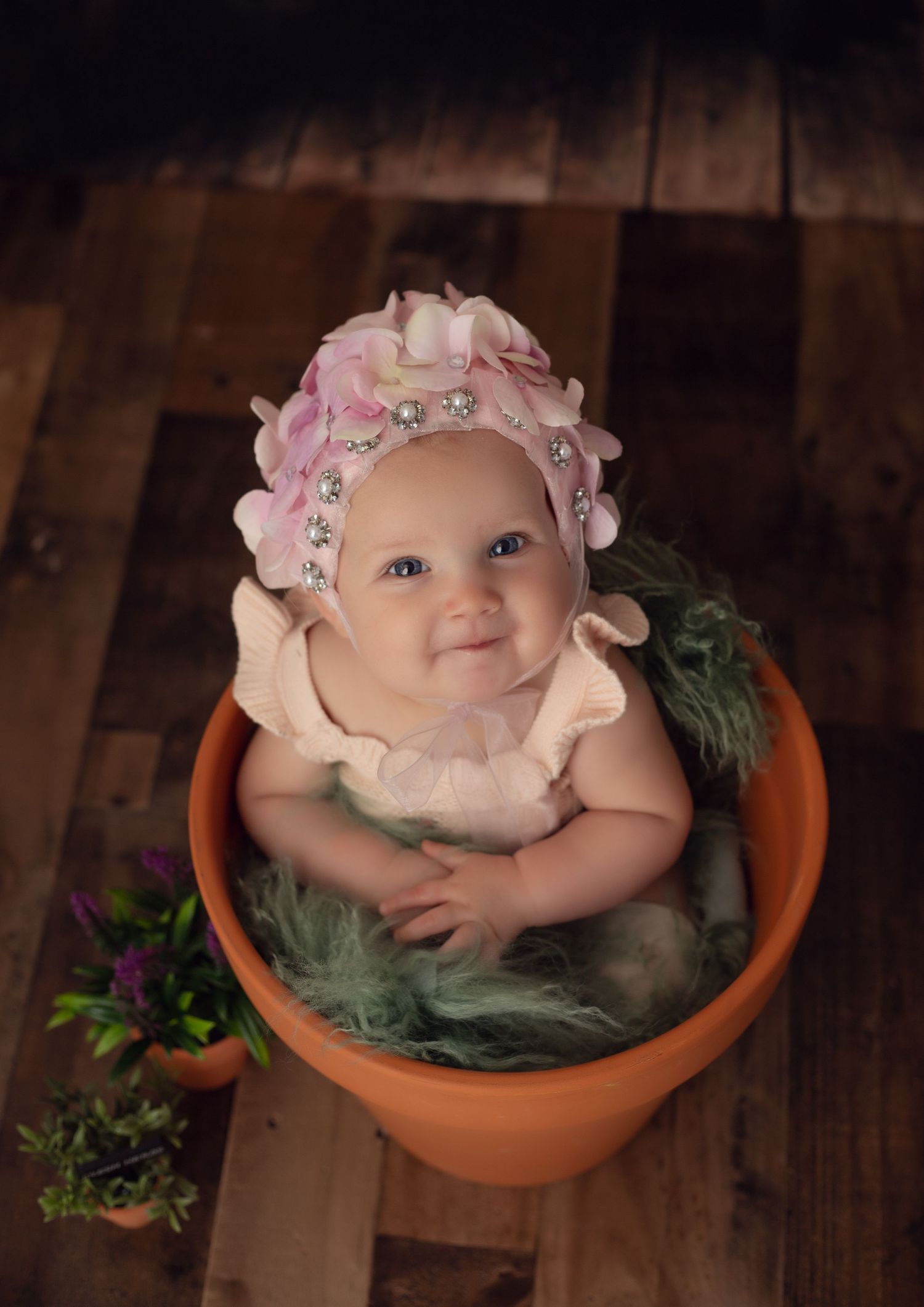 Adorable baby girl sitting in a terracotta flower pot wearing a pink floral bonnet and ruffled outfit on rustic wood floor.
