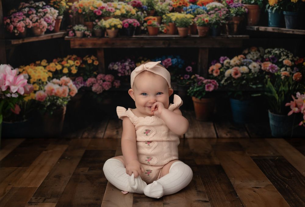 Smiling baby girl in pink romper sitting on wood floor in front of colorful flower shop backdrop.