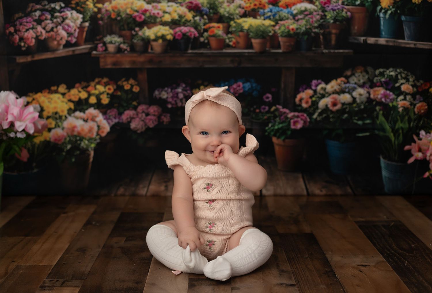 Smiling baby girl in pink romper sitting on wood floor in front of colorful flower shop backdrop.