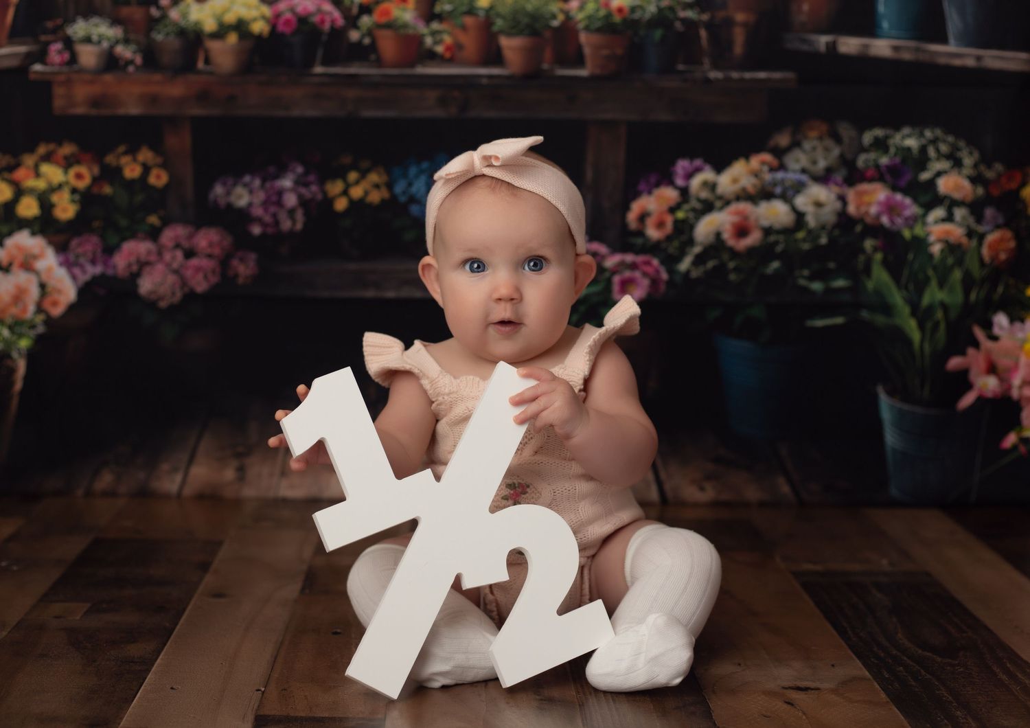 Baby girl in pink romper sitting with a 1/2 birthday sign surrounded by colorful flowers on wooden floor.