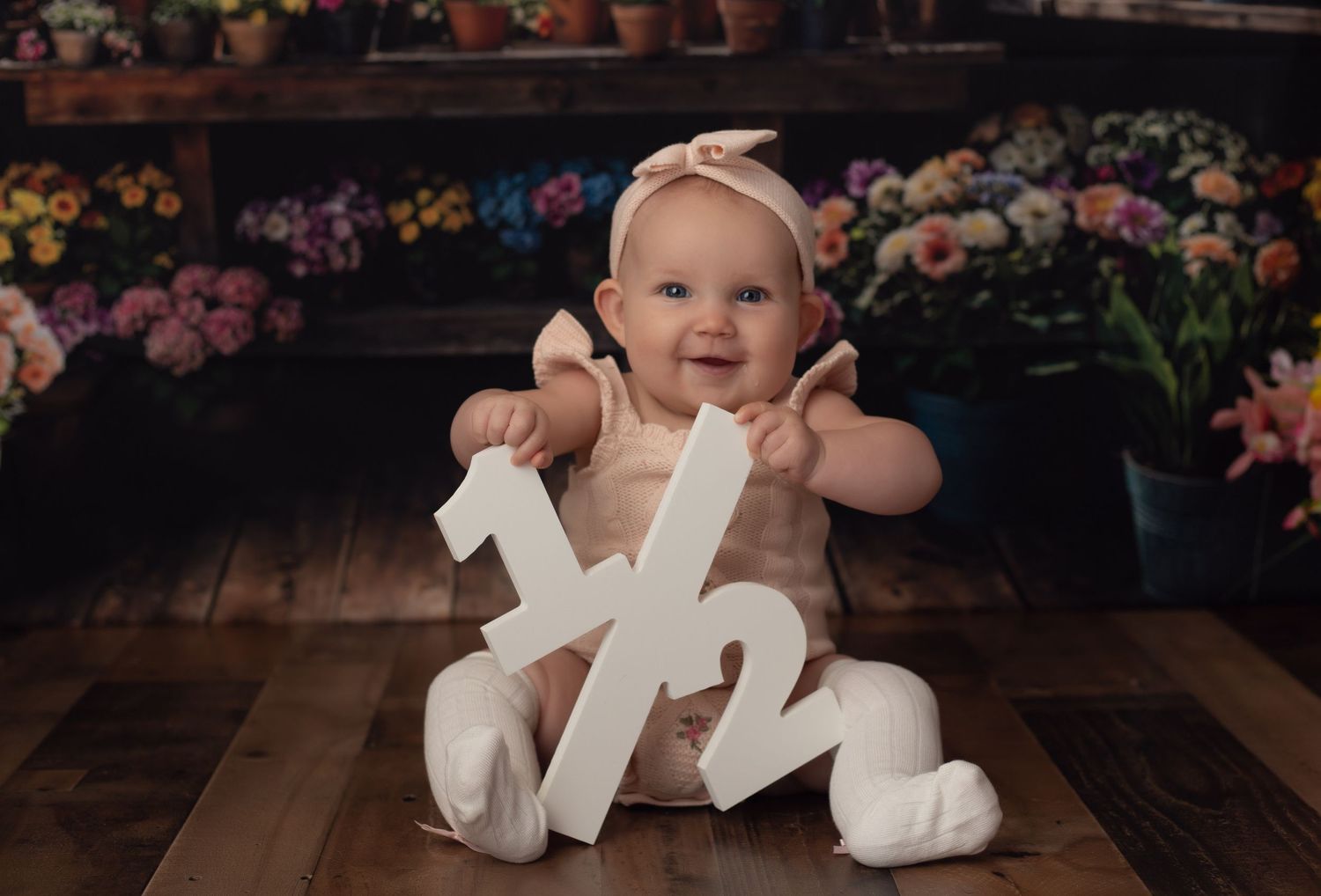 Smiling baby girl in cream outfit holding a 1/2 sign, celebrating her half birthday in a floral studio setting.