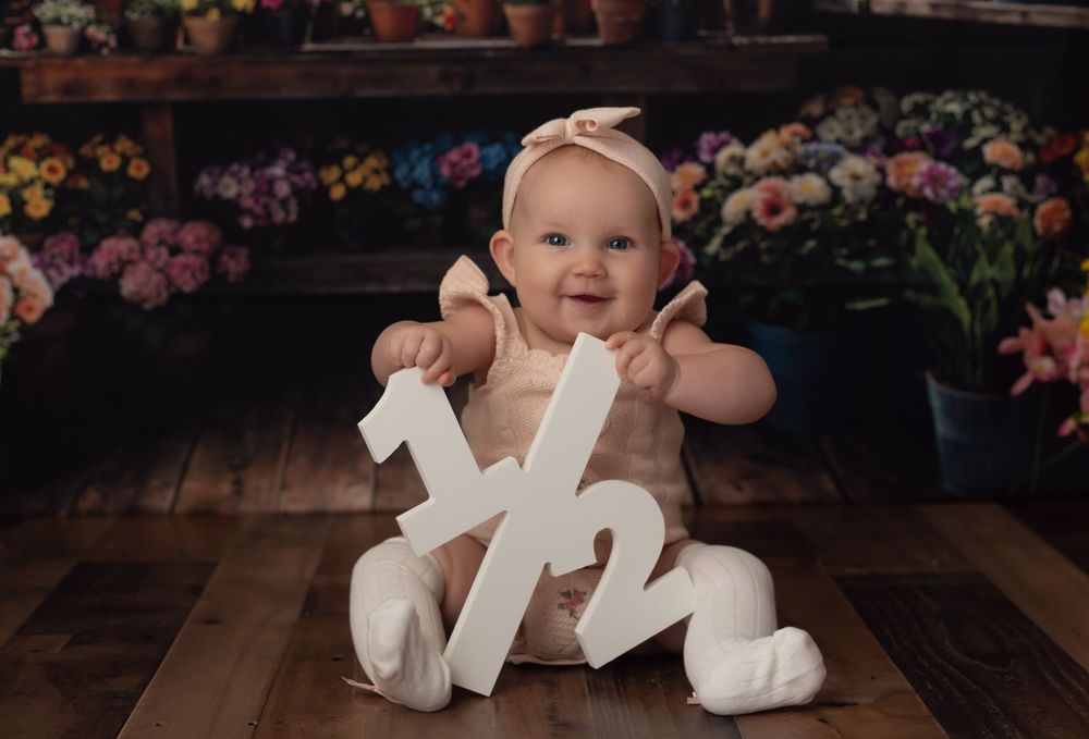 Smiling baby girl in cream outfit holding a 1/2 sign, celebrating her half birthday in a floral studio setting.