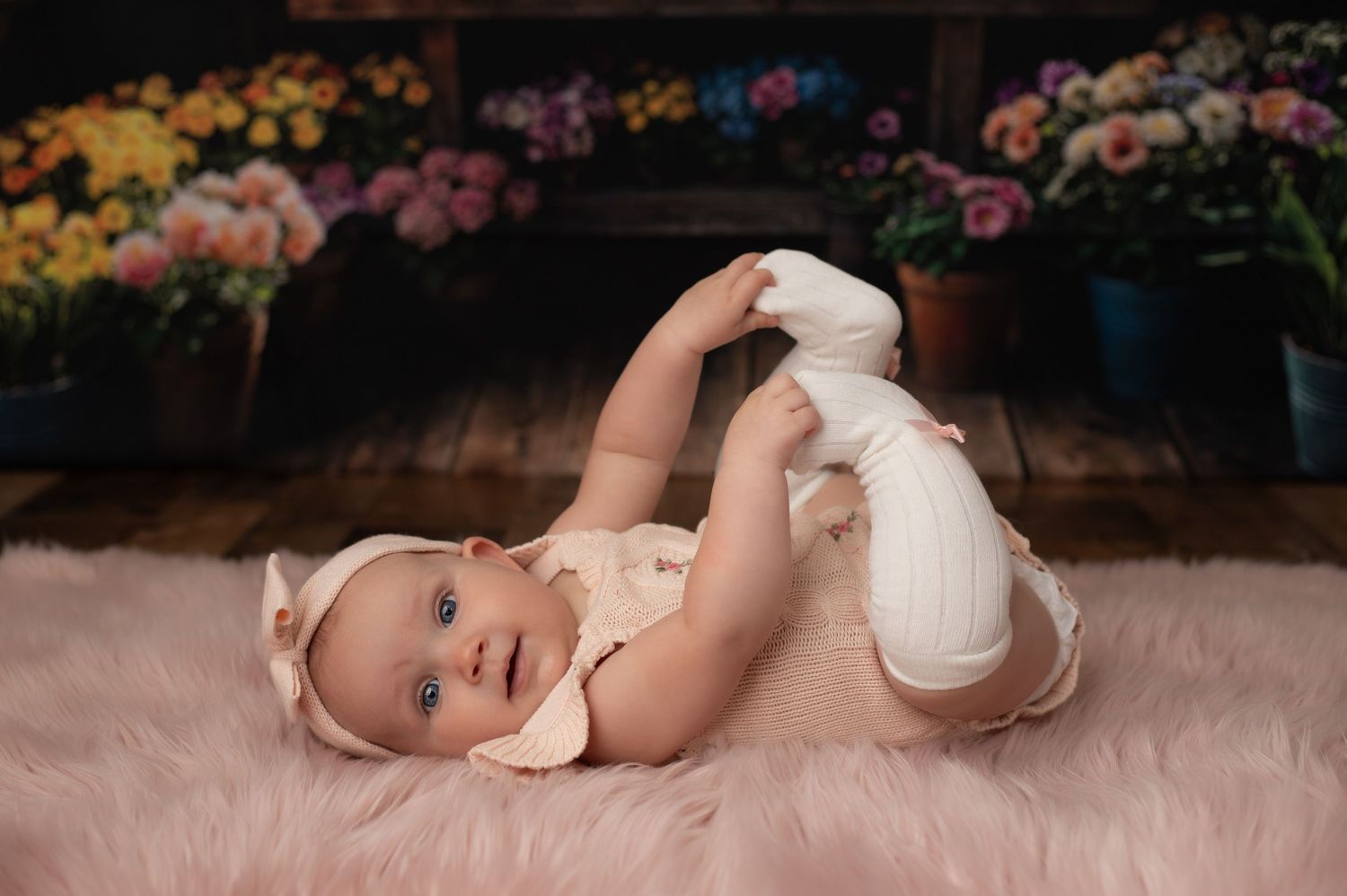 A cute baby in a cream outfit lies on a pink rug, holding her feet up, with colorful flowers in the background.