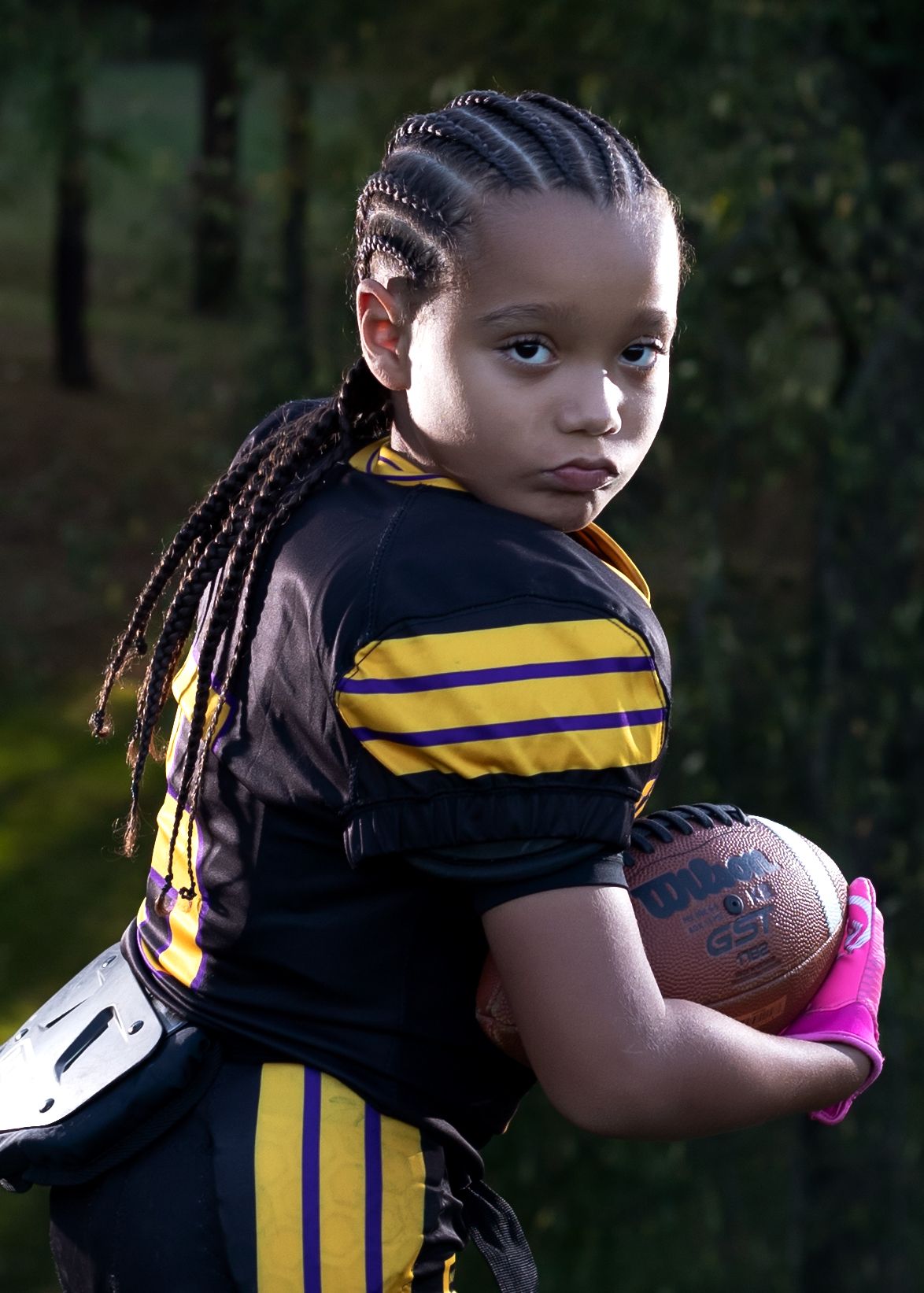 Young football player in black and gold uniform holding a Wilson football, wearing pink gloves, with braided hair.