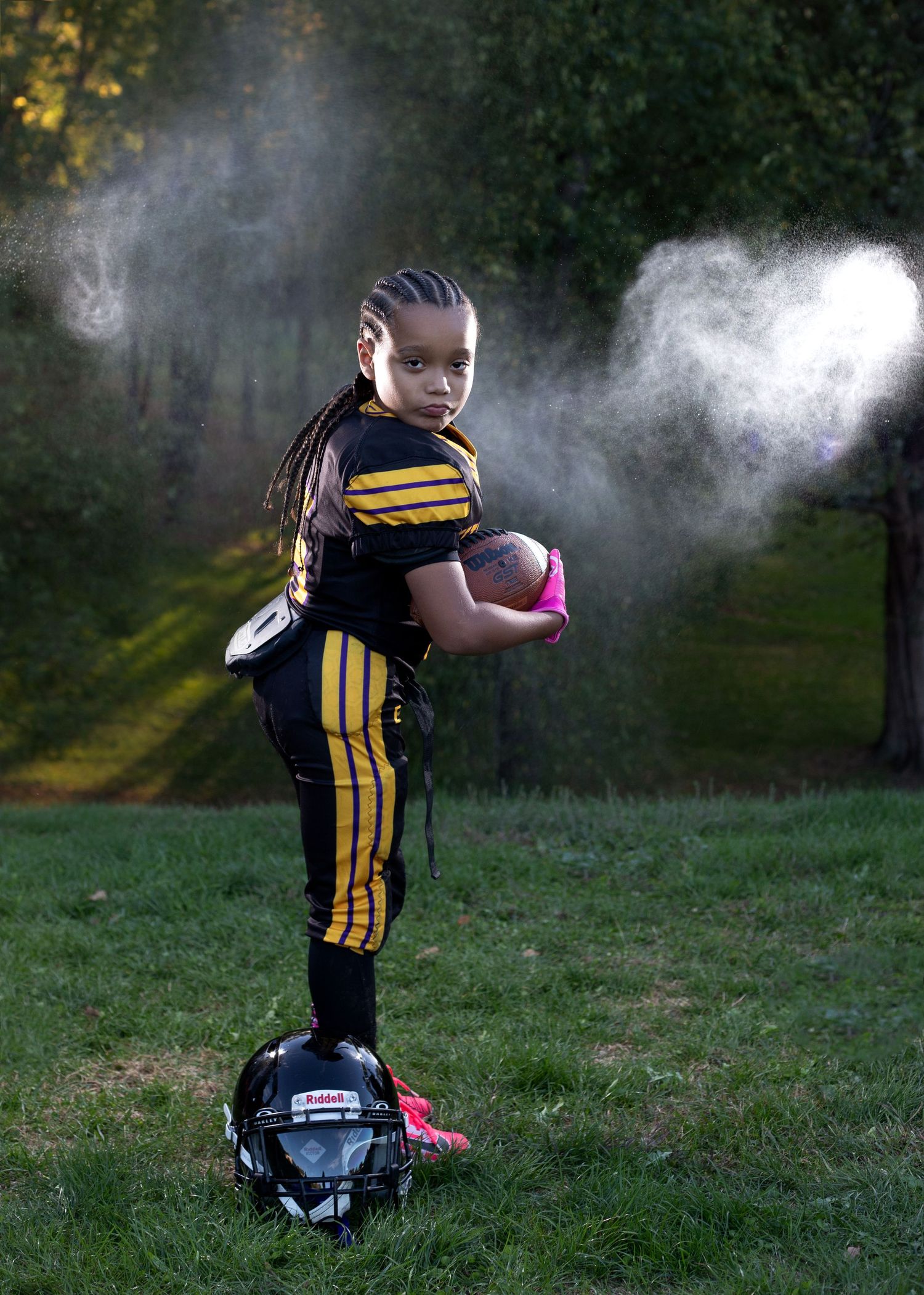 Young girl in black and gold football uniform holds ball confidently, helmet at feet, mist in background.