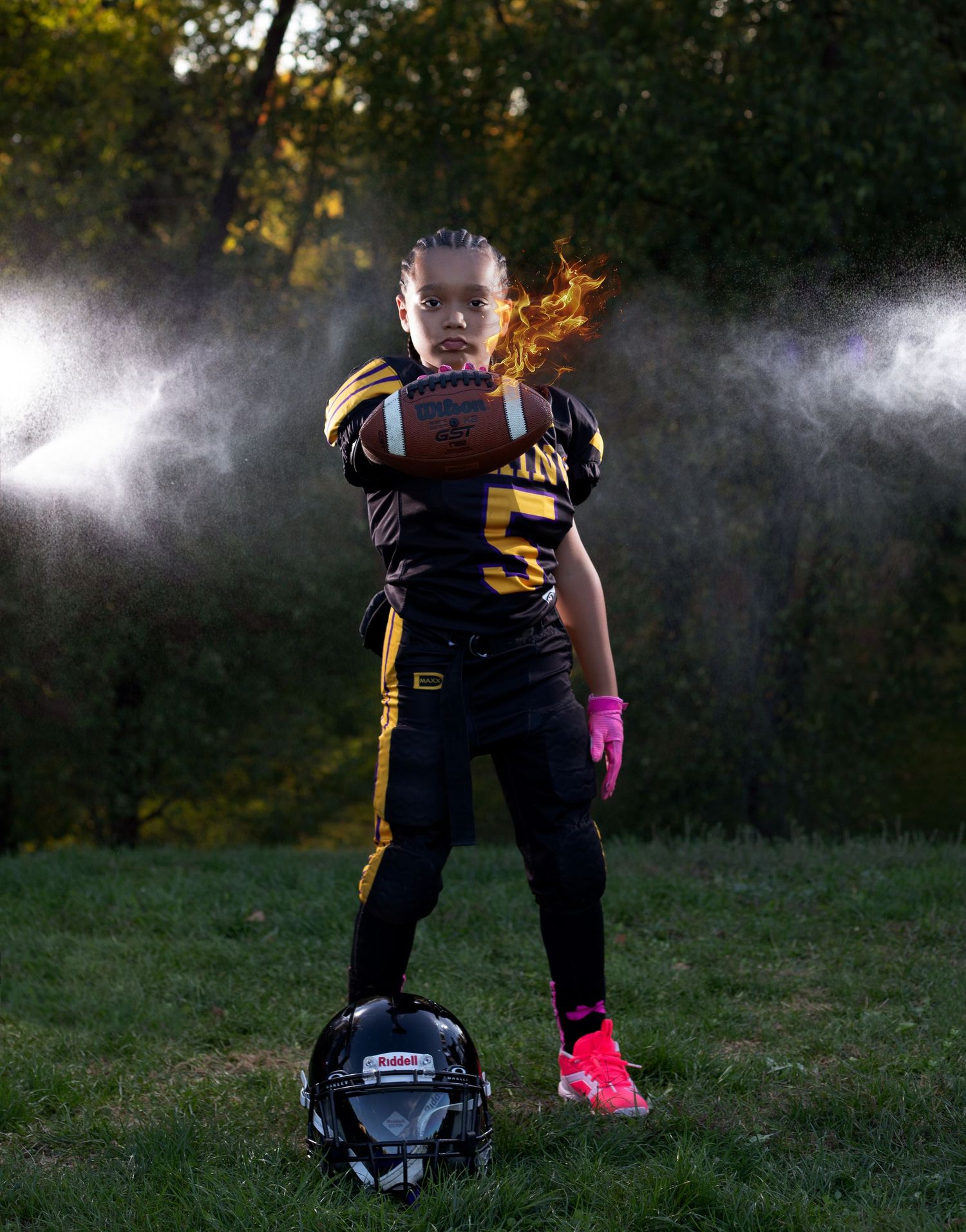 Young girl in black and gold football uniform #5 holds ball confidently, pink cleats, helmet at feet, dramatic backlit outdoor setting.