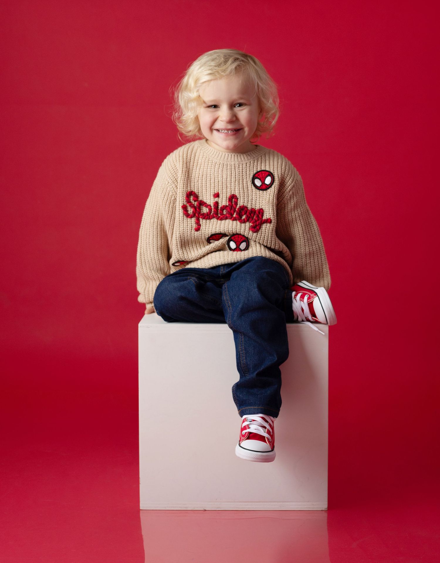 Toddler boy wearing a beige Spidey knit sweater, dark jeans, and red sneakers, sitting on a white cube against a red background.