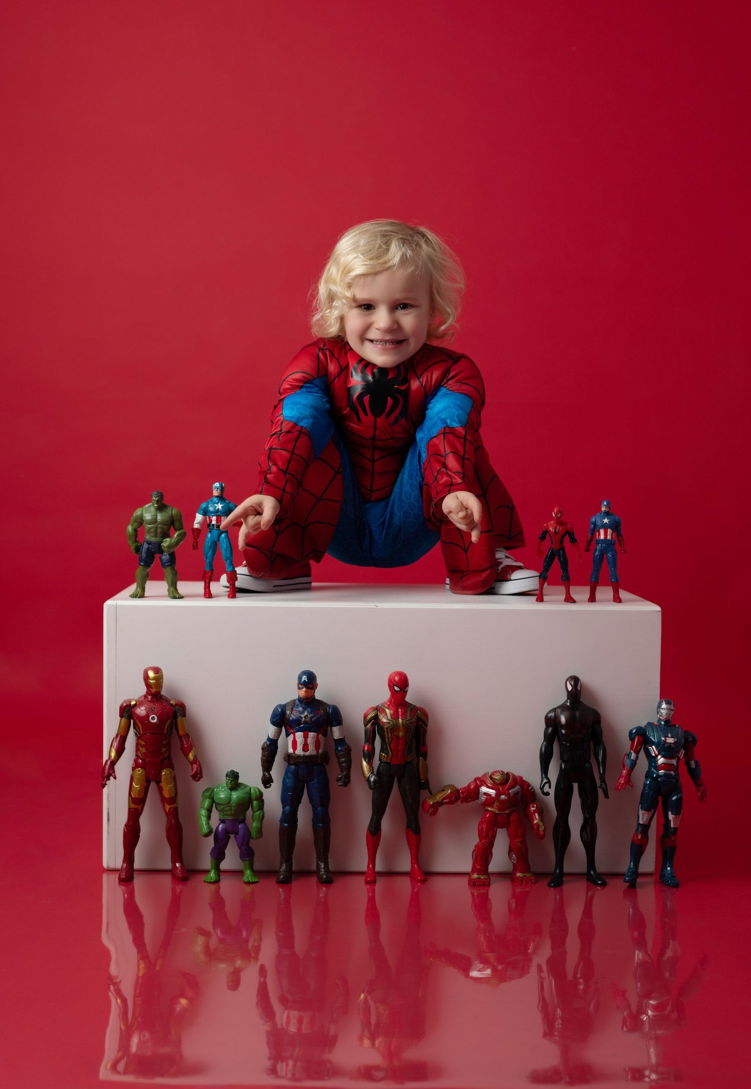 Smiling child in Spider-Man costume poses with Marvel superhero action figures on white block against red background.