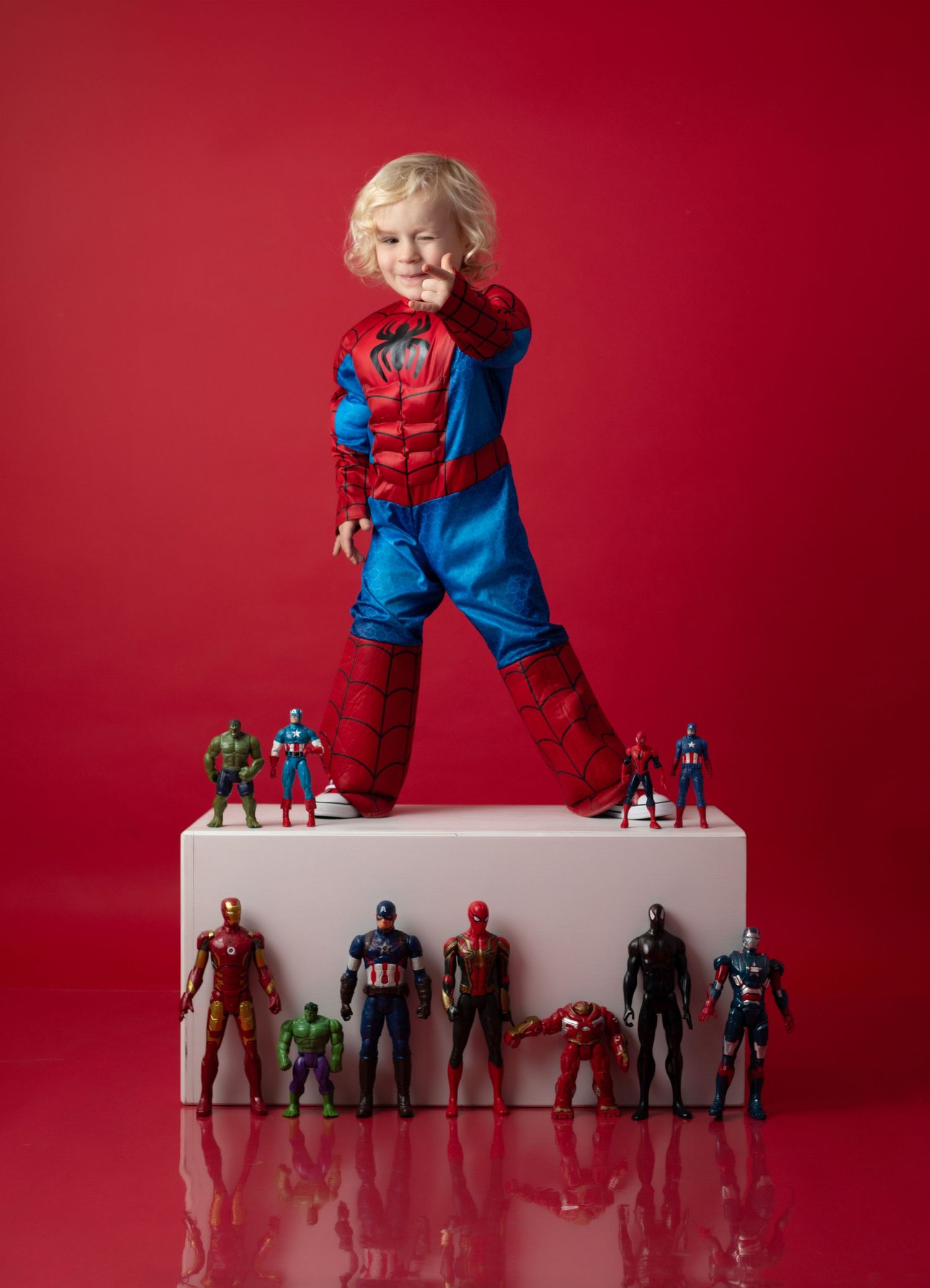 A young child dressed in a Spider-Man costume stands on a white podium surrounded by Marvel superhero action figures on a red background.