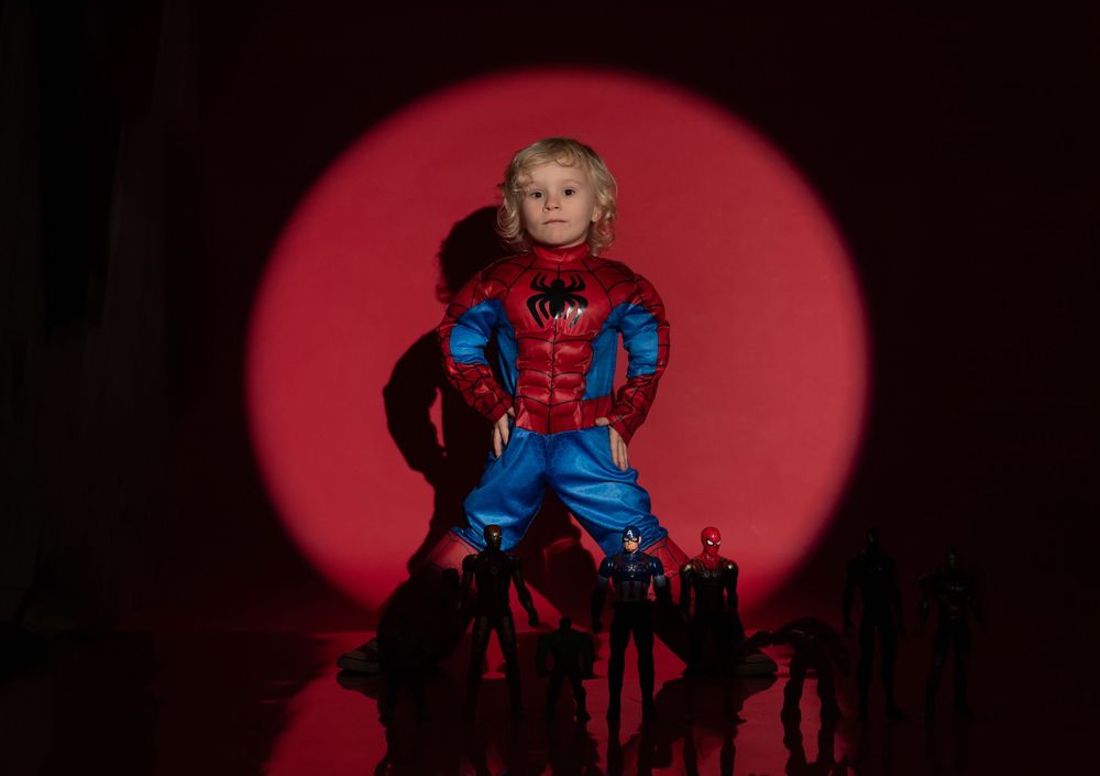 Young child in Spider-Man costume posed heroically under a red spotlight with action figures.