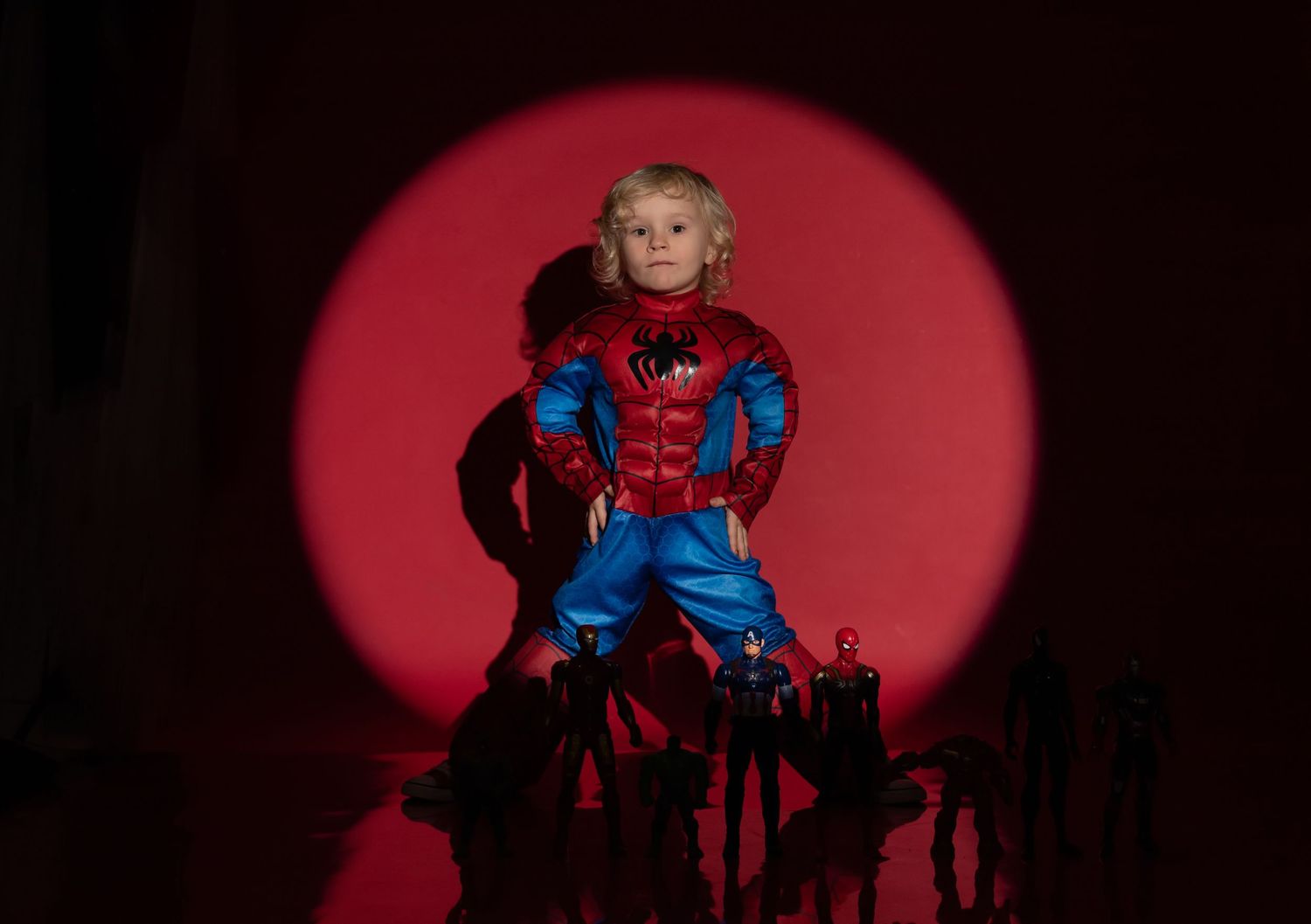 Young child in Spider-Man costume posed heroically under a red spotlight with action figures.