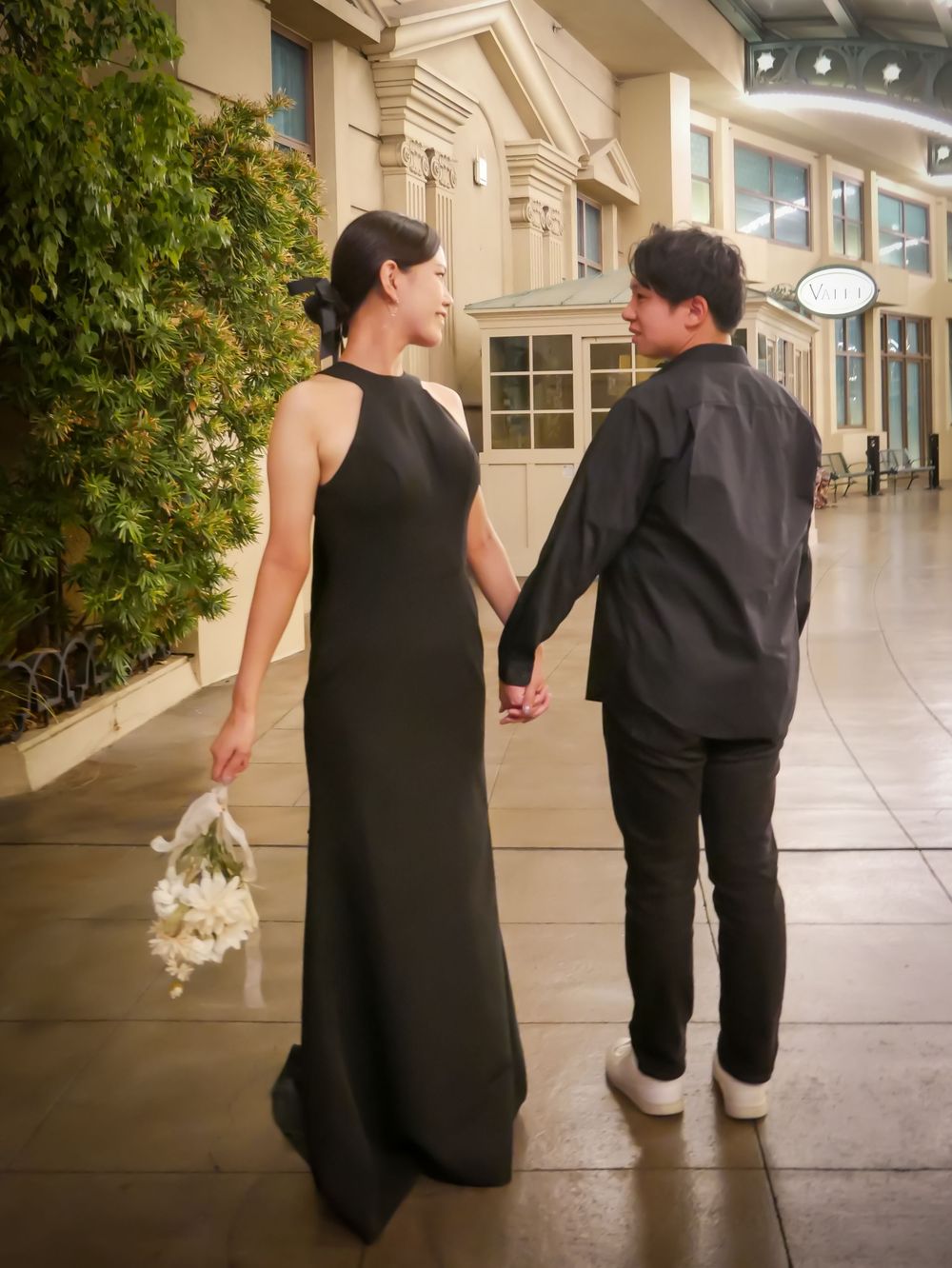 Couple in matching black outfits holding hands, woman in elegant halter gown holding white bouquet, walking together.