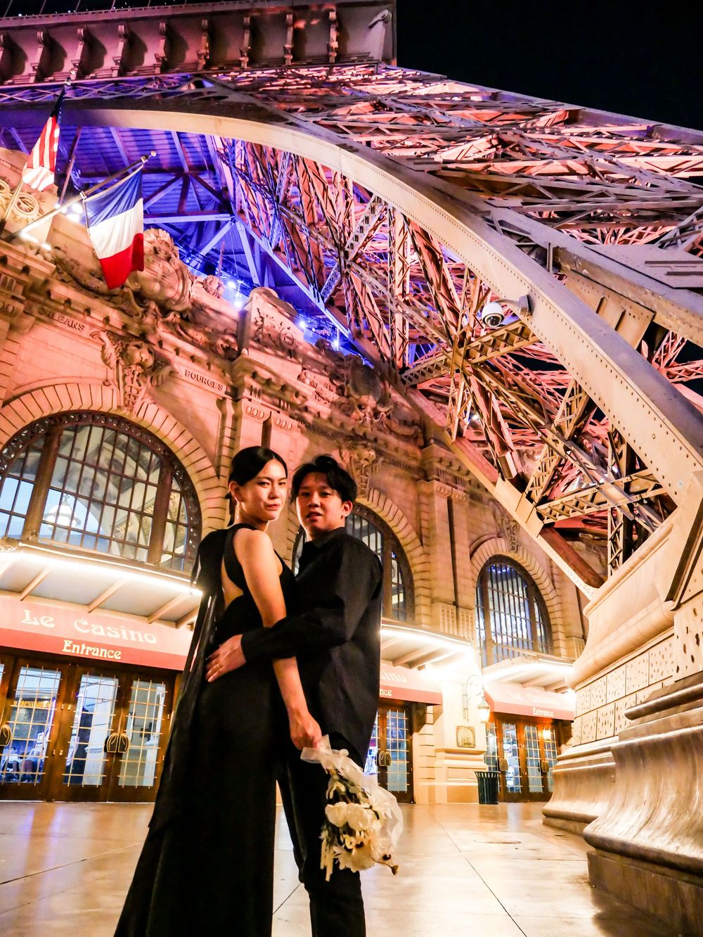Couple in black formal attire poses romantically under the illuminated Eiffel Tower at night in Paris.