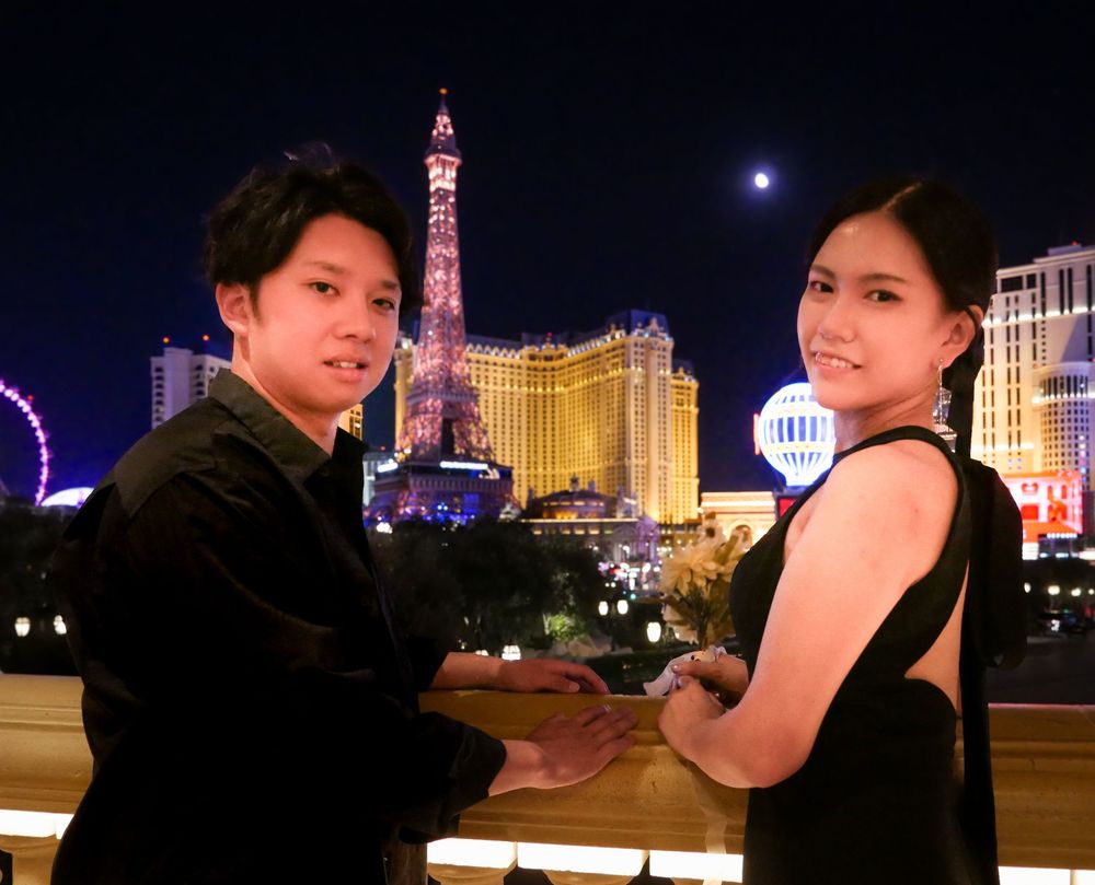 Couple holding hands on Las Vegas Strip balcony at night with Eiffel Tower replica and city lights behind them.