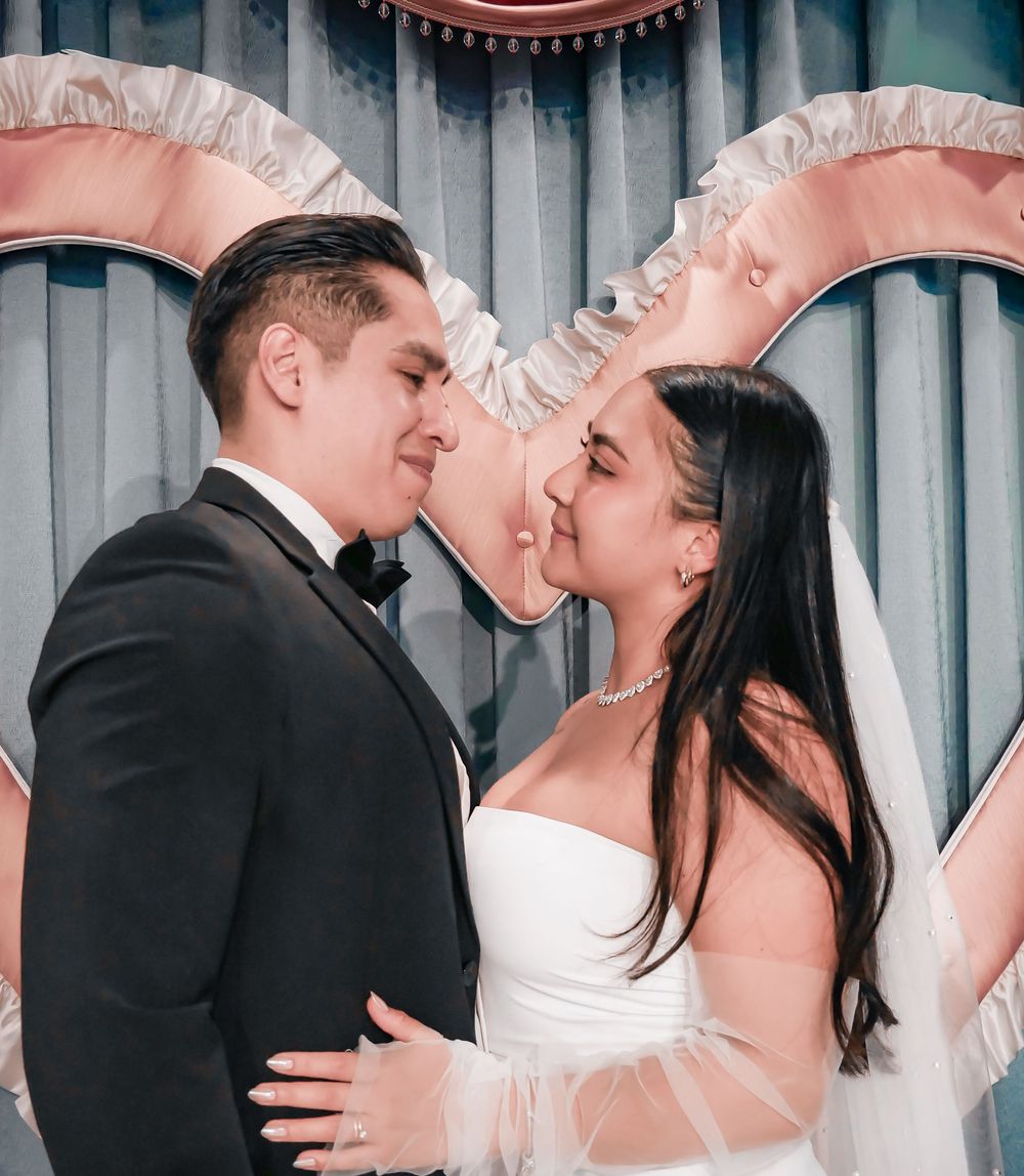A bride and groom share a romantic moment, nearly kissing, dressed in wedding attire before a decorative backdrop.