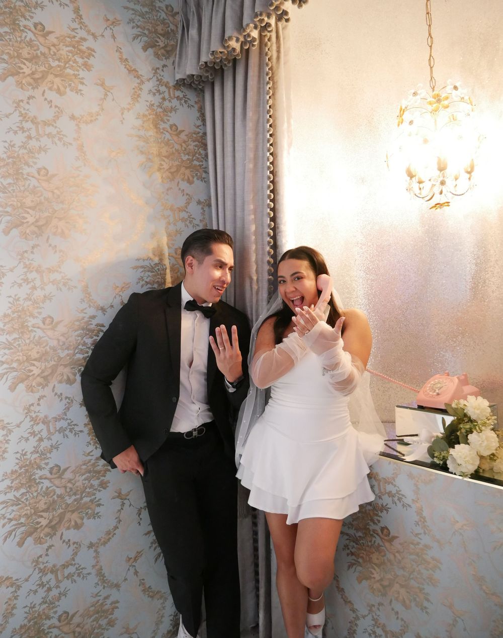 Newlywed couple in wedding attire showing off rings, man in tuxedo and woman in short white dress with veil.
