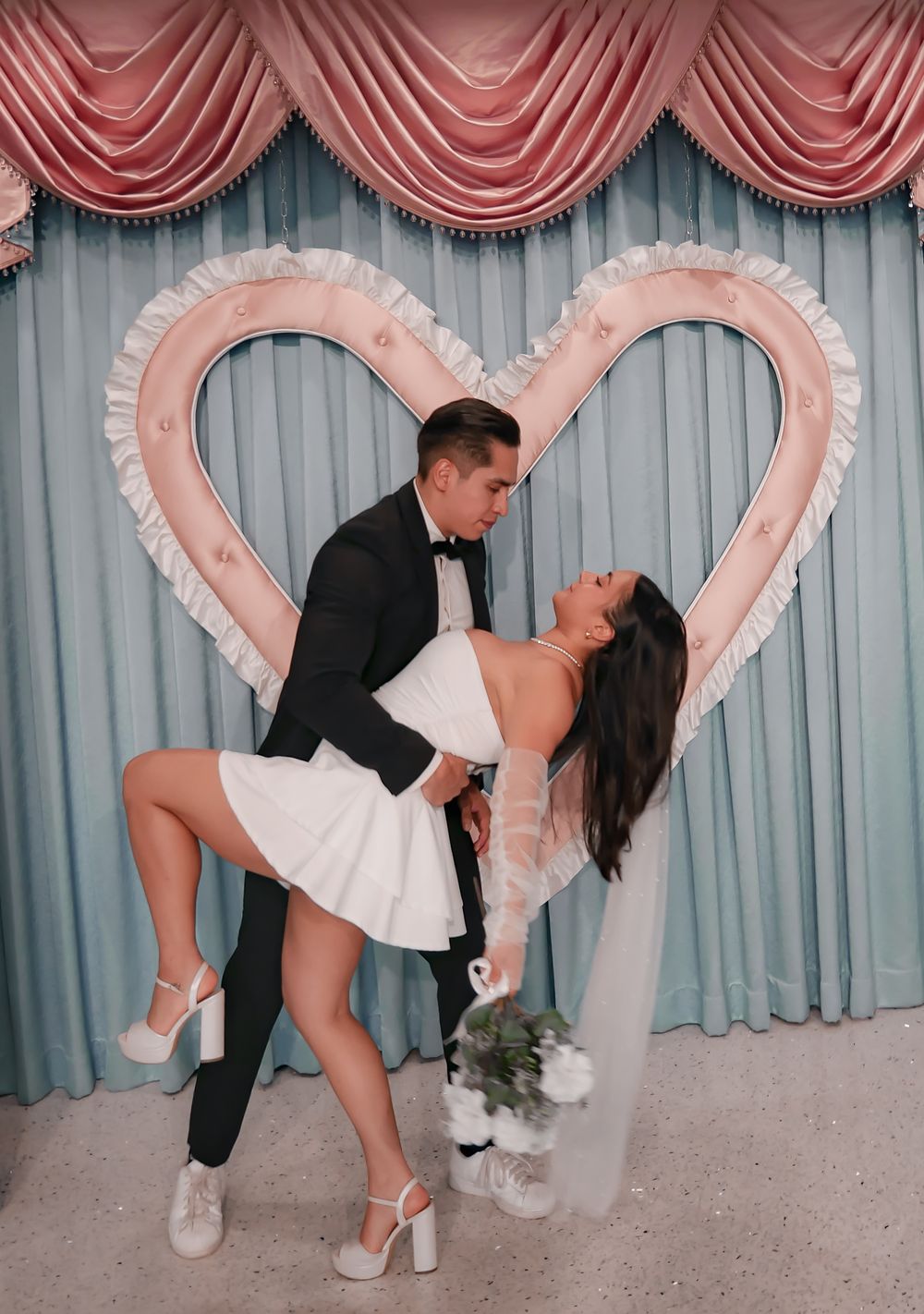 Bride and groom share a romantic dip kiss in front of a pink heart backdrop at a Las Vegas wedding chapel.