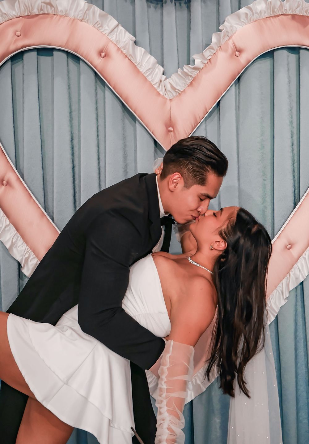 Couple sharing a romantic kiss in wedding attire in front of a large pink heart backdrop.
