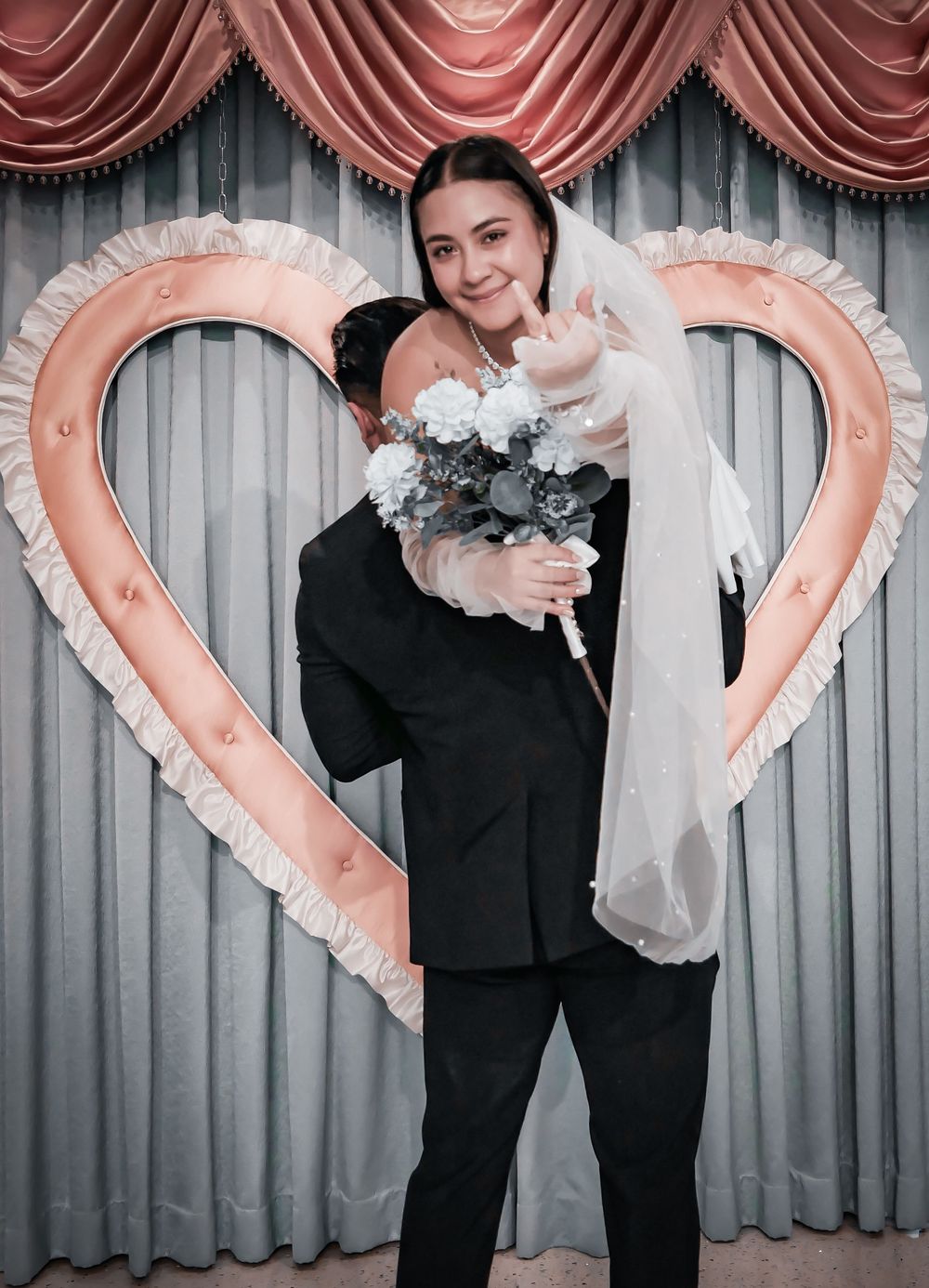 Bride in white veil holding flower bouquet embraced by groom in black suit before heart-shaped decoration.