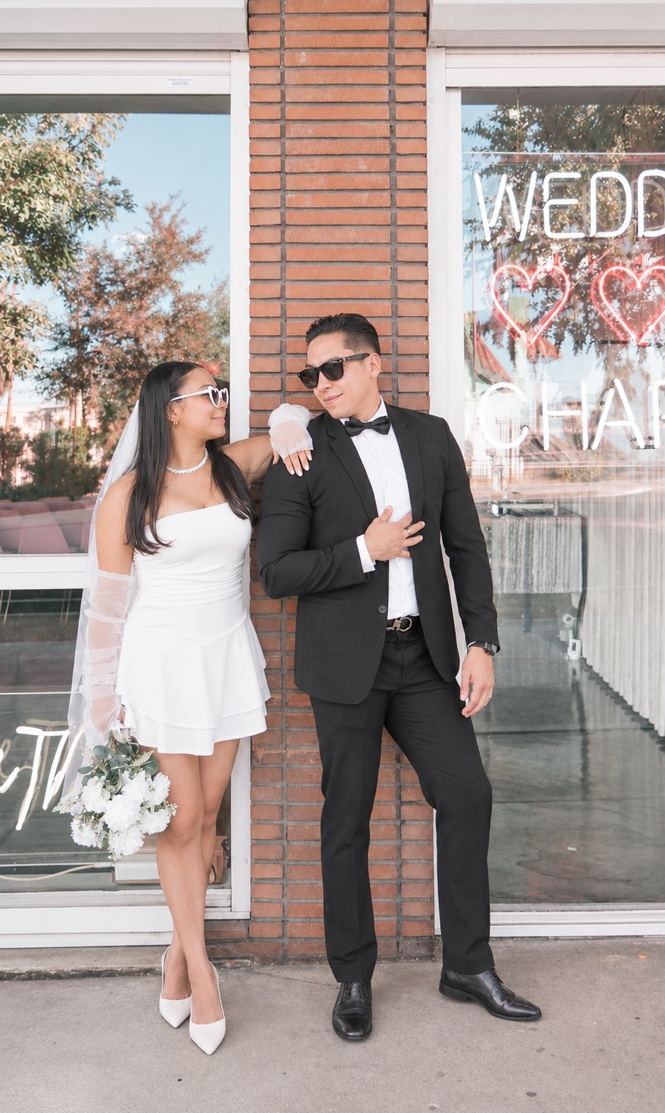 Bride in white mini dress and groom in black tuxedo posing together outside a wedding chapel.