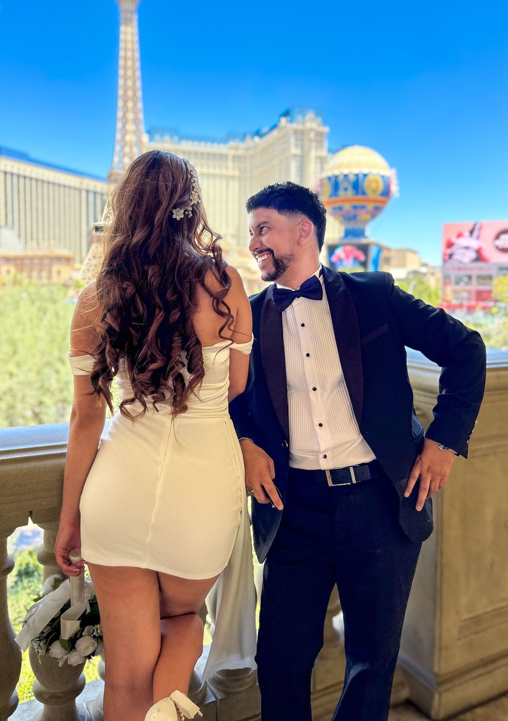 Bride in white mini dress and groom in tuxedo smiling together with Paris Las Vegas Eiffel Tower behind them.