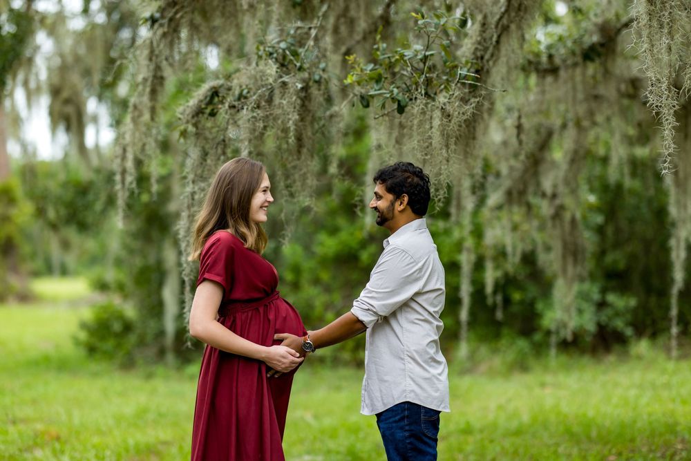 Pregnant woman in red dress holds hands with man outdoors under moss-draped trees in a maternity photo session.