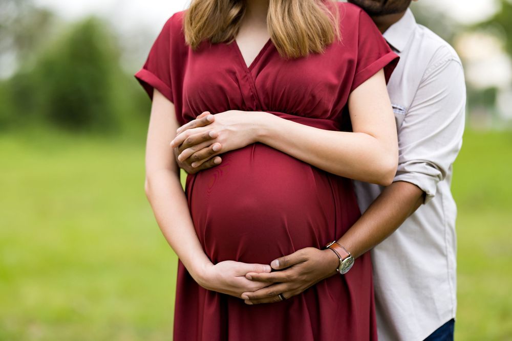 Pregnant woman in red dress with partner embracing her baby bump outdoors in a green field.