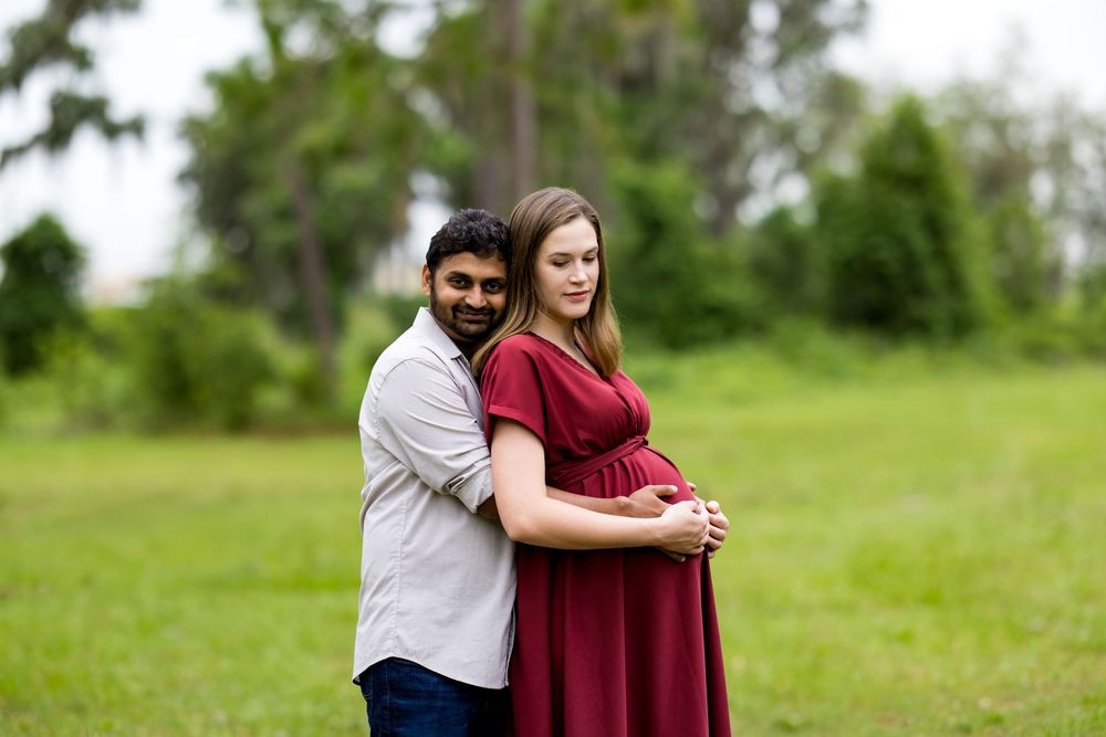 Couple poses outdoors on green grass, pregnant woman in red dress cradled by partner during maternity photo session.