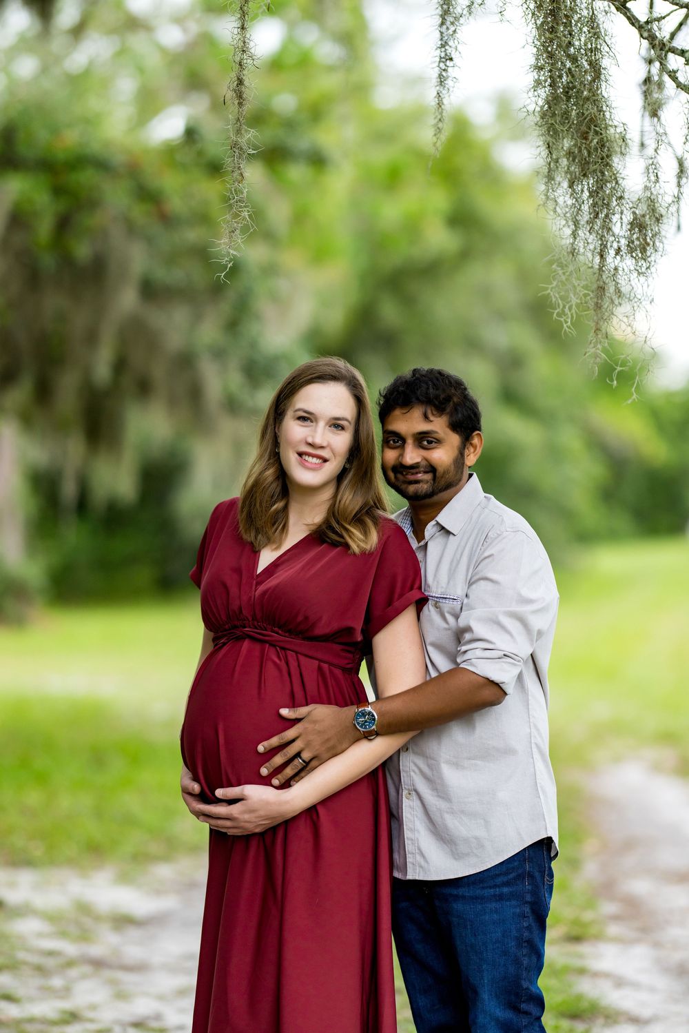 Pregnant couple posing outdoors under Spanish moss trees, woman in red maternity dress, man in gray shirt holding her belly.