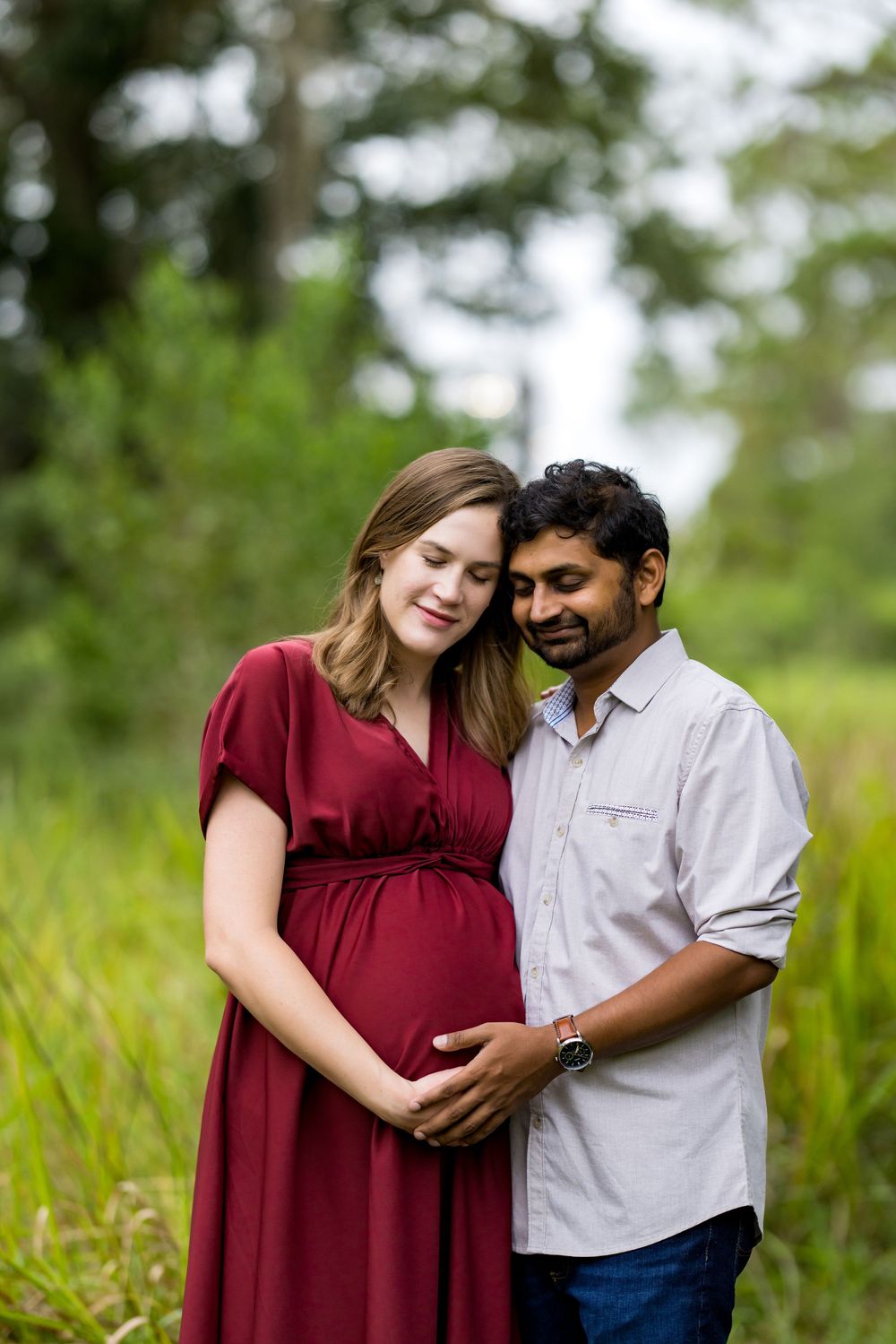 Interracial couple sharing tender maternity moment outdoors, pregnant woman in red dress with partner embracing her baby bump.