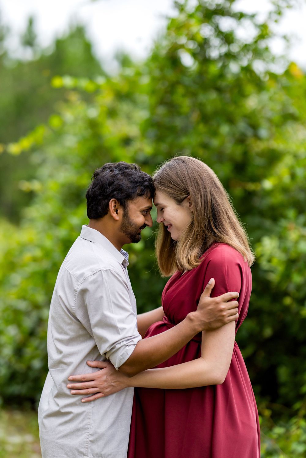 A couple embracing outdoors, smiling at each other with lush green trees in the background.