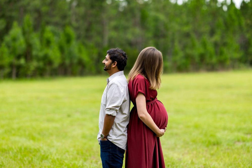 Pregnant couple standing back-to-back in a green field, woman in red dress cradling baby bump during maternity photoshoot.