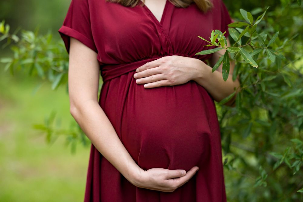 Pregnant woman in red dress cradling baby bump outdoors near green foliage.
