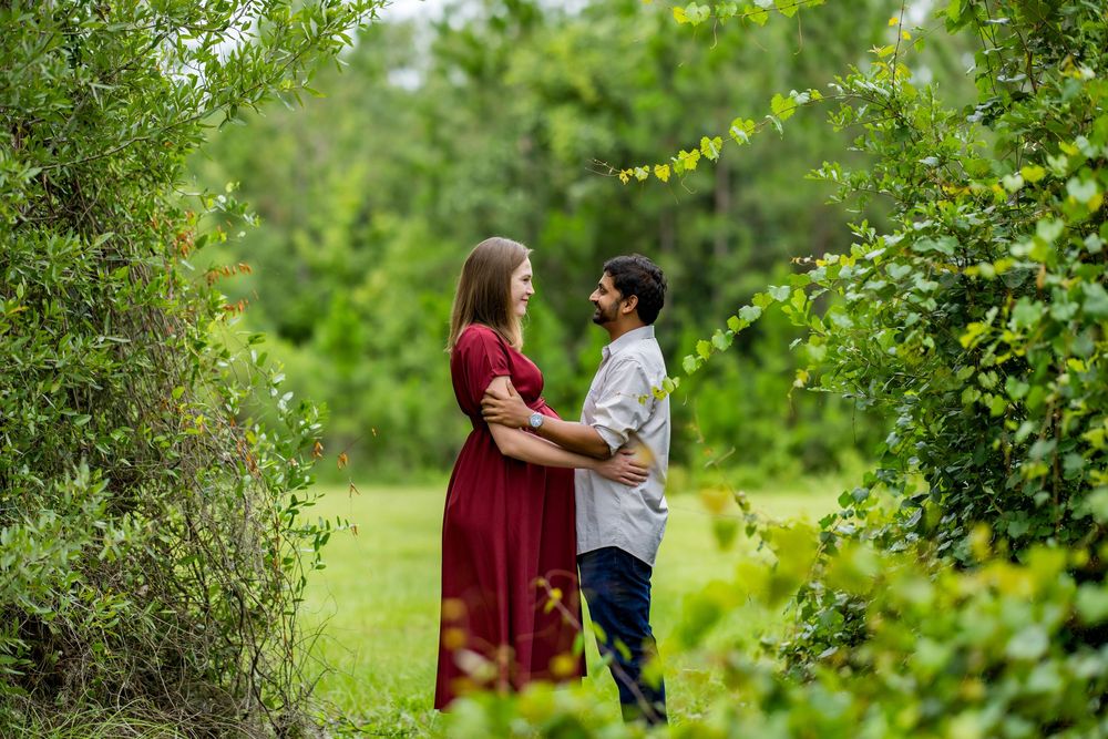 Couple embracing in lush green garden, woman in red dress and man in casual attire, romantic outdoor engagement photo.
