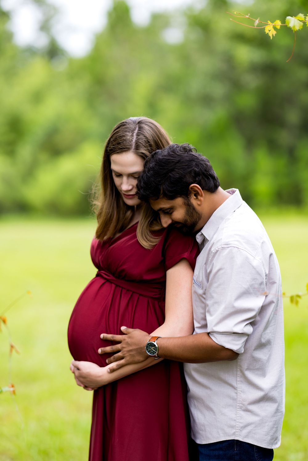 Couple sharing a tender moment outdoors, pregnant woman in red dress with partner embracing her baby bump in a green park.