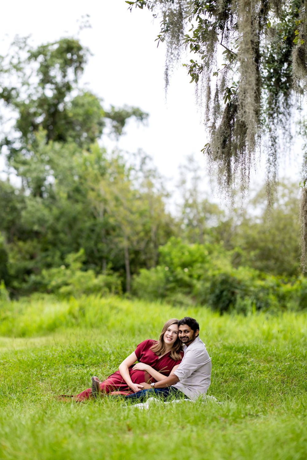Couple sitting together on lush green grass under Spanish moss-draped tree, woman in red dress leaning on man in white shirt.