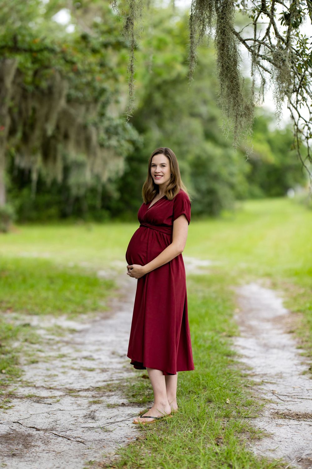 Pregnant woman in red dress standing on a rustic path surrounded by lush greenery and Spanish moss.