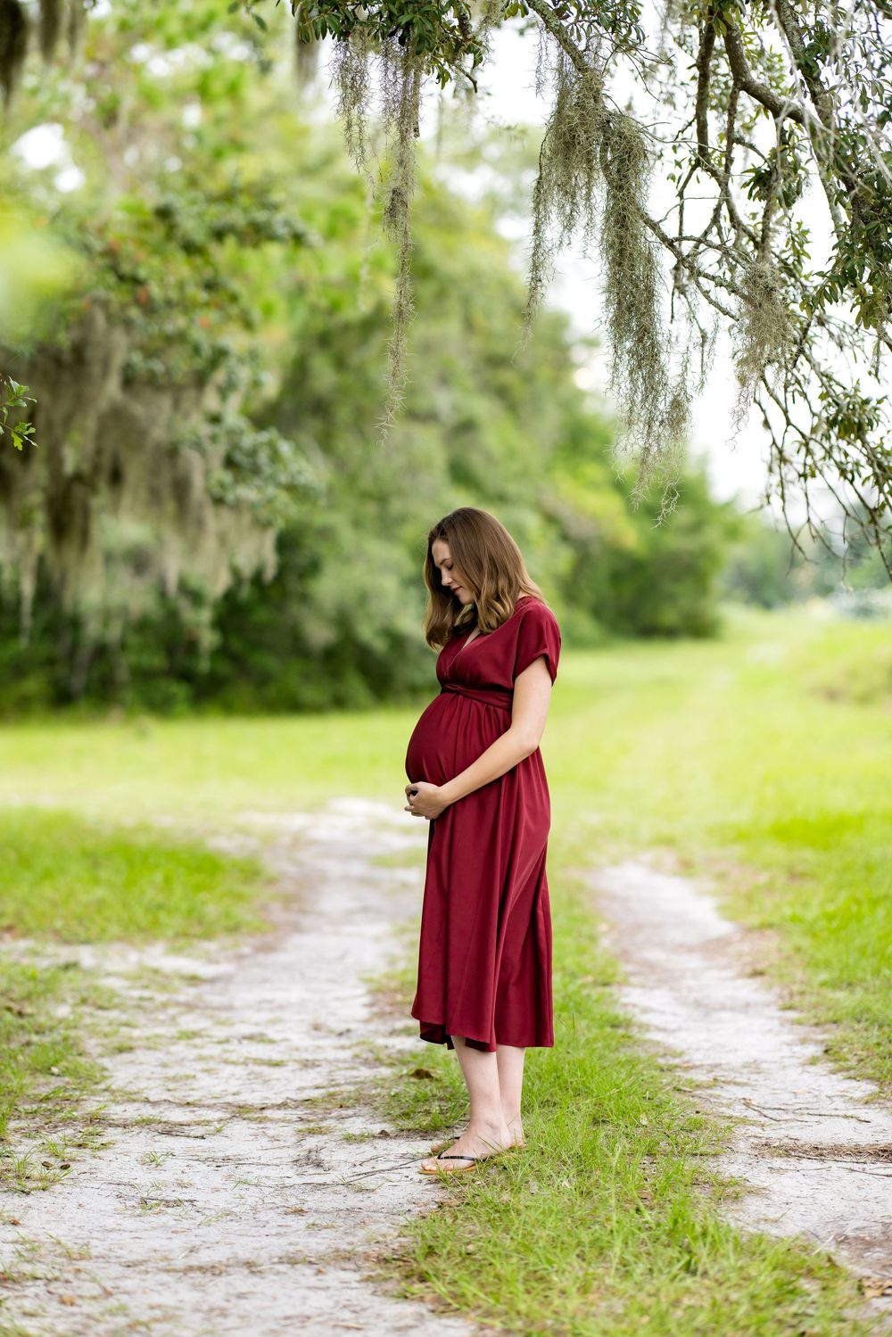 Pregnant woman in red dress standing on a path under moss-draped trees, cradling her baby bump outdoors.
